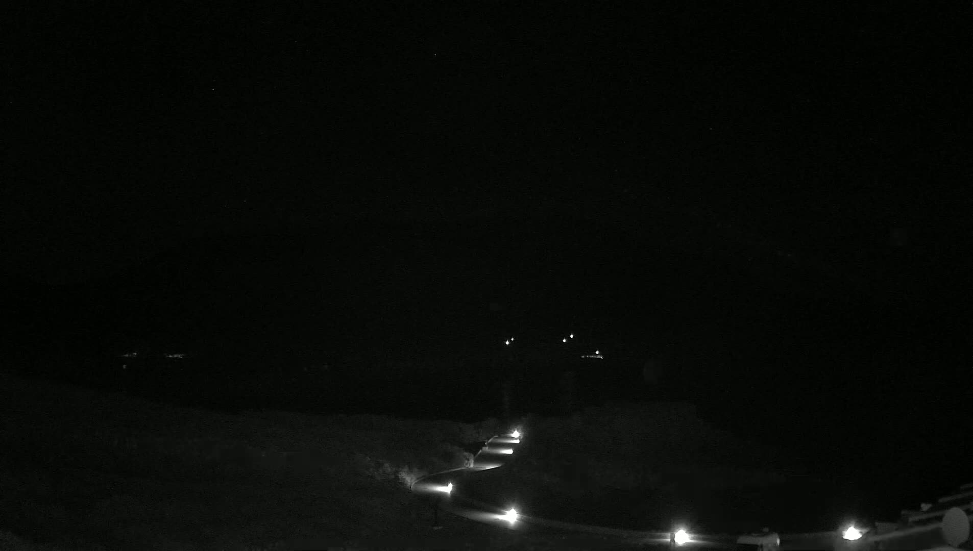 A dark, nighttime scene shows a winding road lit by spaced-out lamps, curving through a sparsely vegetated landscape under a clear sky.
