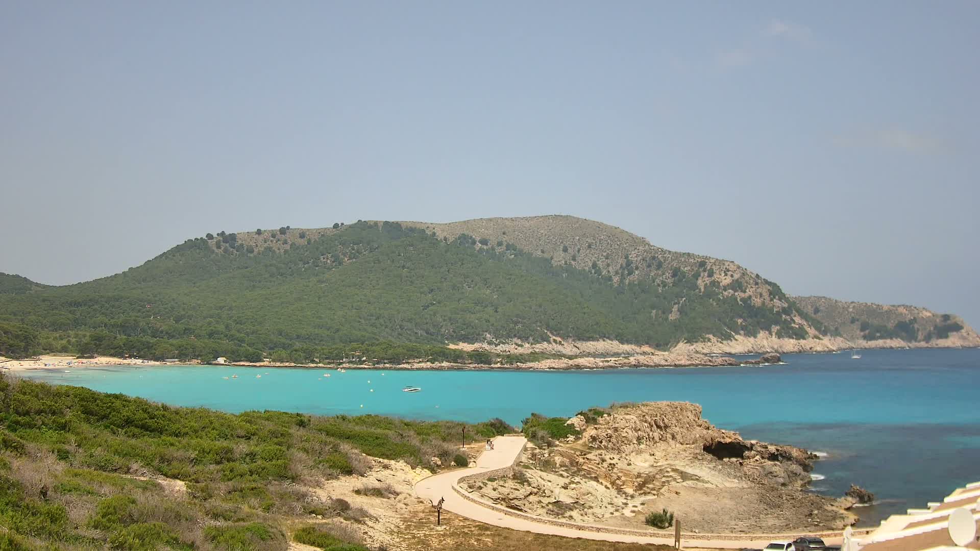 A winding road leads to a turquoise bay with a sandy beach, nestled beneath a green, rocky hill on a clear, sunny day.