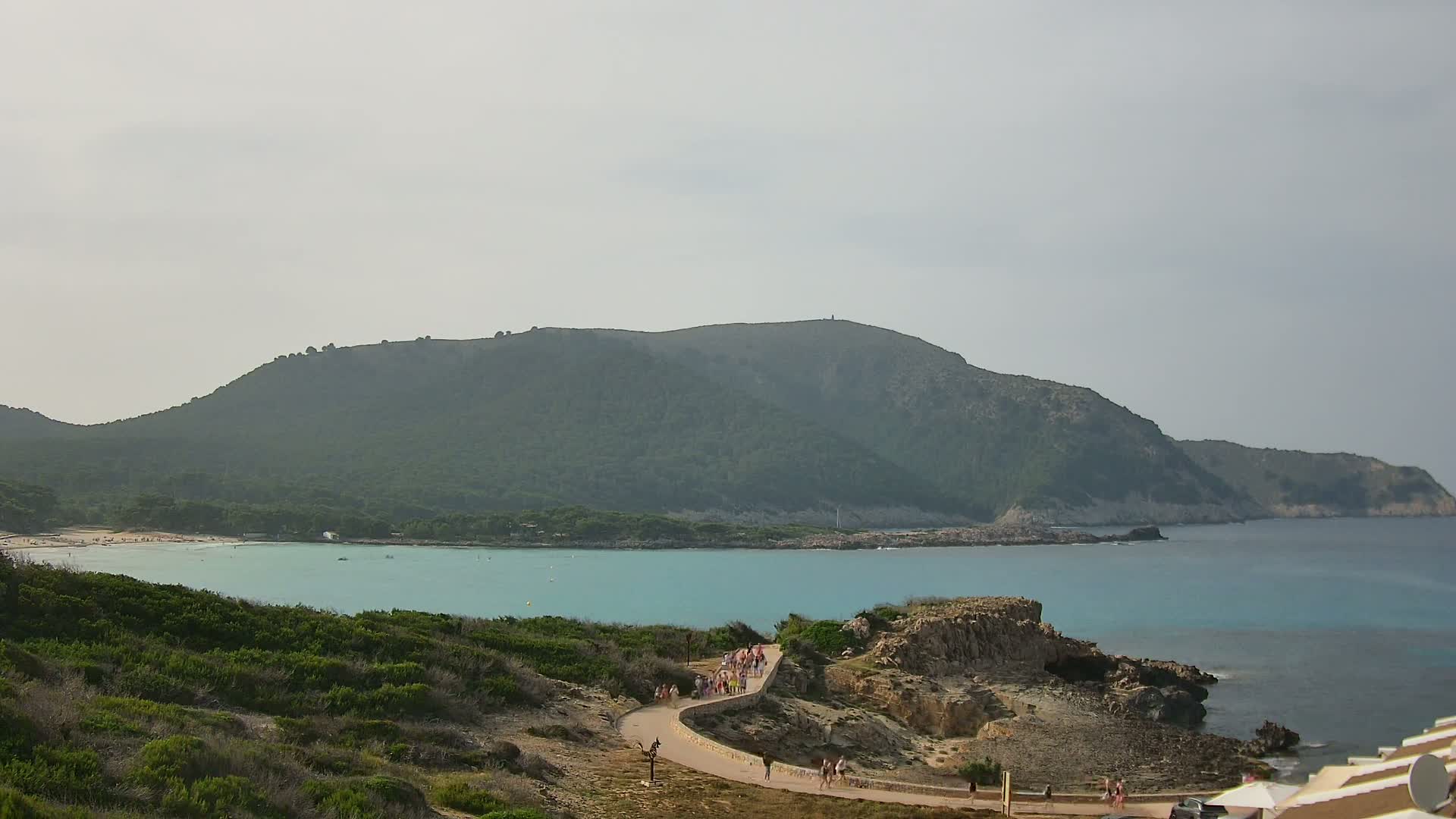 A coastal scene under a cloudy sky shows a turquoise bay with a sandy beach, a winding path with people walking on it, and a large, green hill in the background.