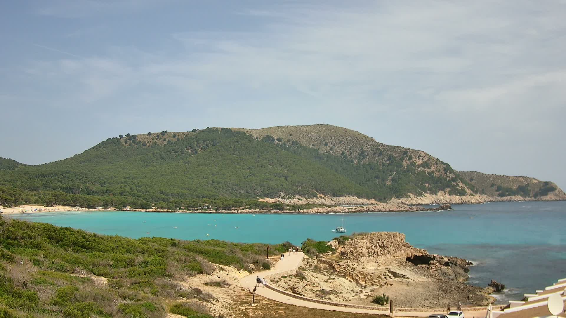 A path winds along a rocky coastline overlooking a calm, turquoise bay with a sandy beach and a green, tree-covered hill in the background under a mostly clear sky.