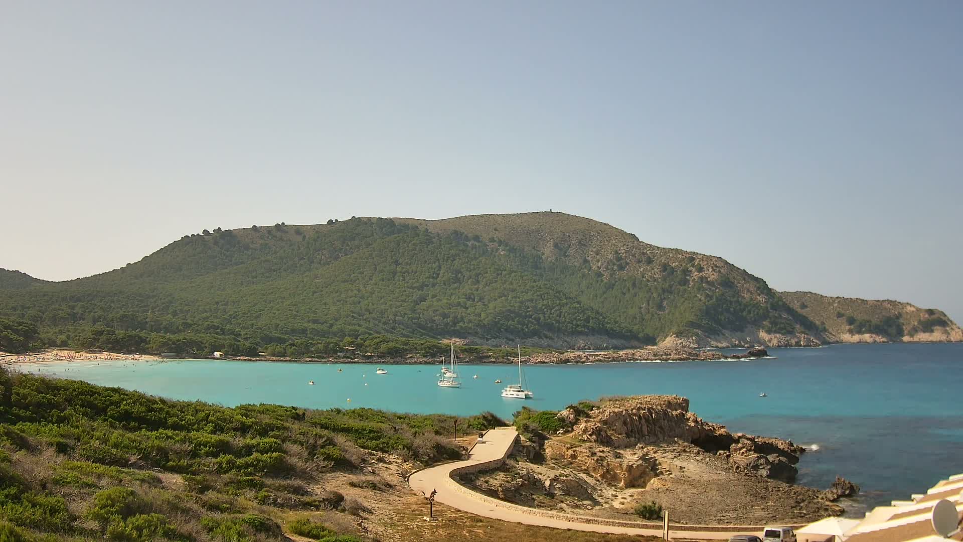 A clear, sunny day reveals a tranquil cove with turquoise water, a sandy beach filled with people, several sailboats, and a verdant, tree-covered hill in the background.