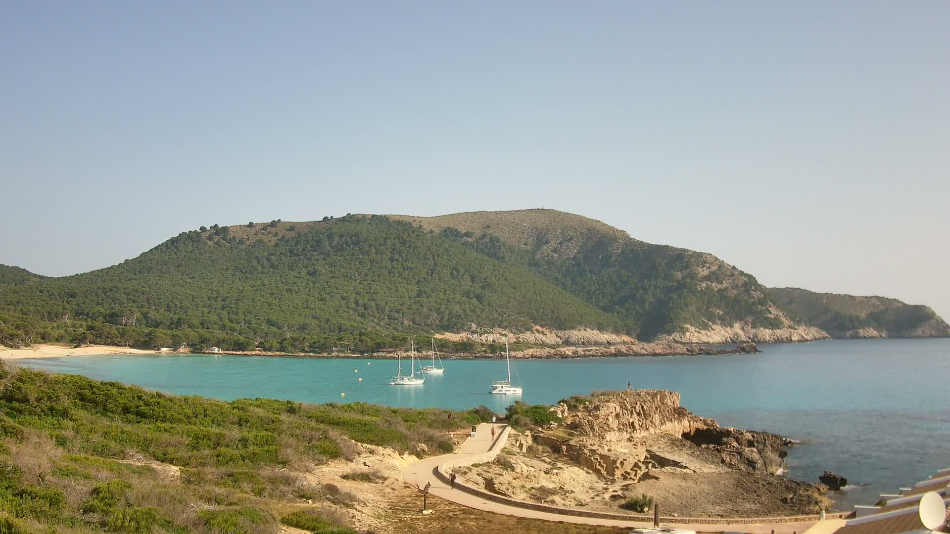 A secluded cove with turquoise water, a sandy beach, and several sailboats is nestled beneath a verdant, hilly coastline on a clear, sunny day.