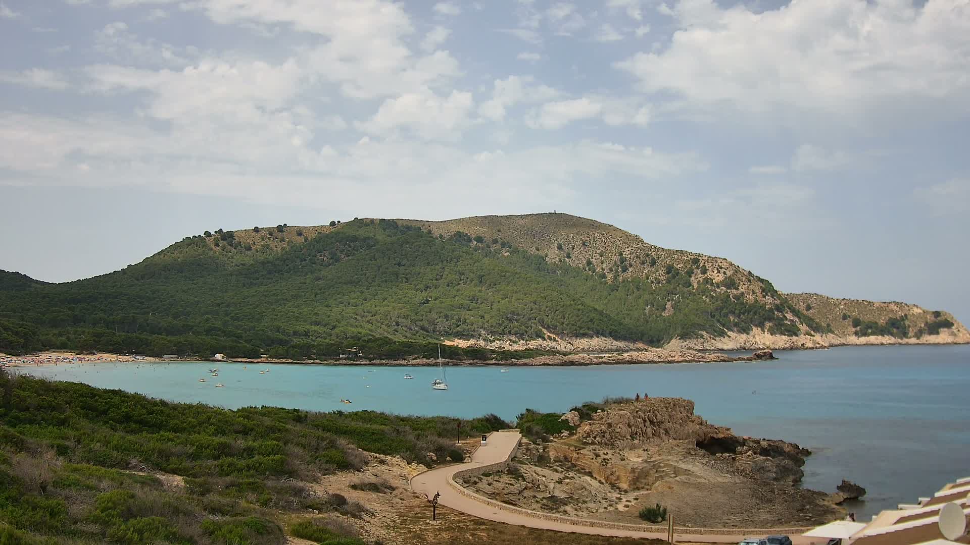A partly cloudy day shows a sandy beach and calm turquoise water curving around a green, rocky hill with a paved path winding through the scrubby landscape.
