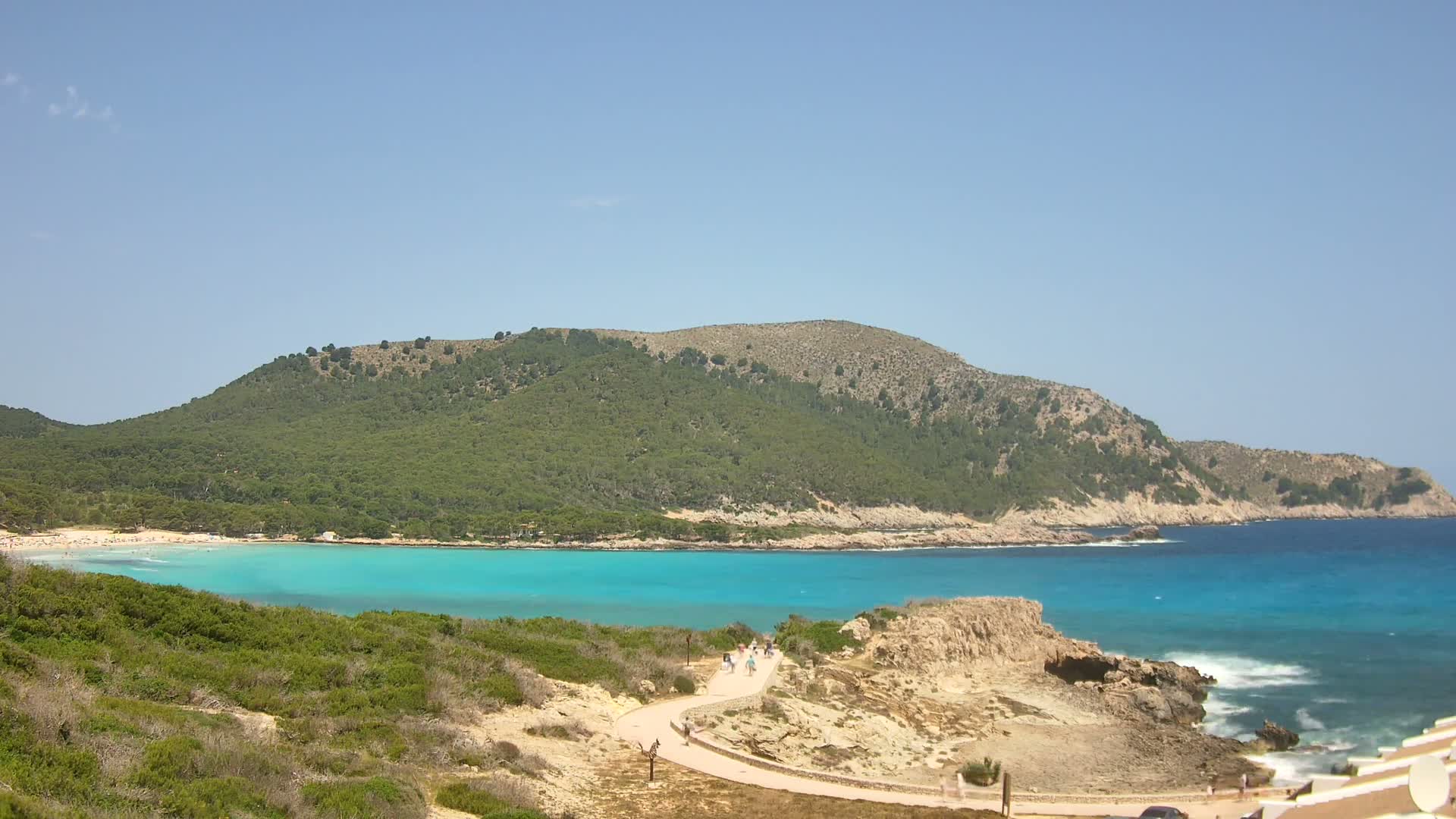 A path winds through scrubby vegetation toward a turquoise bay, backed by a verdant hill under a clear blue sky.