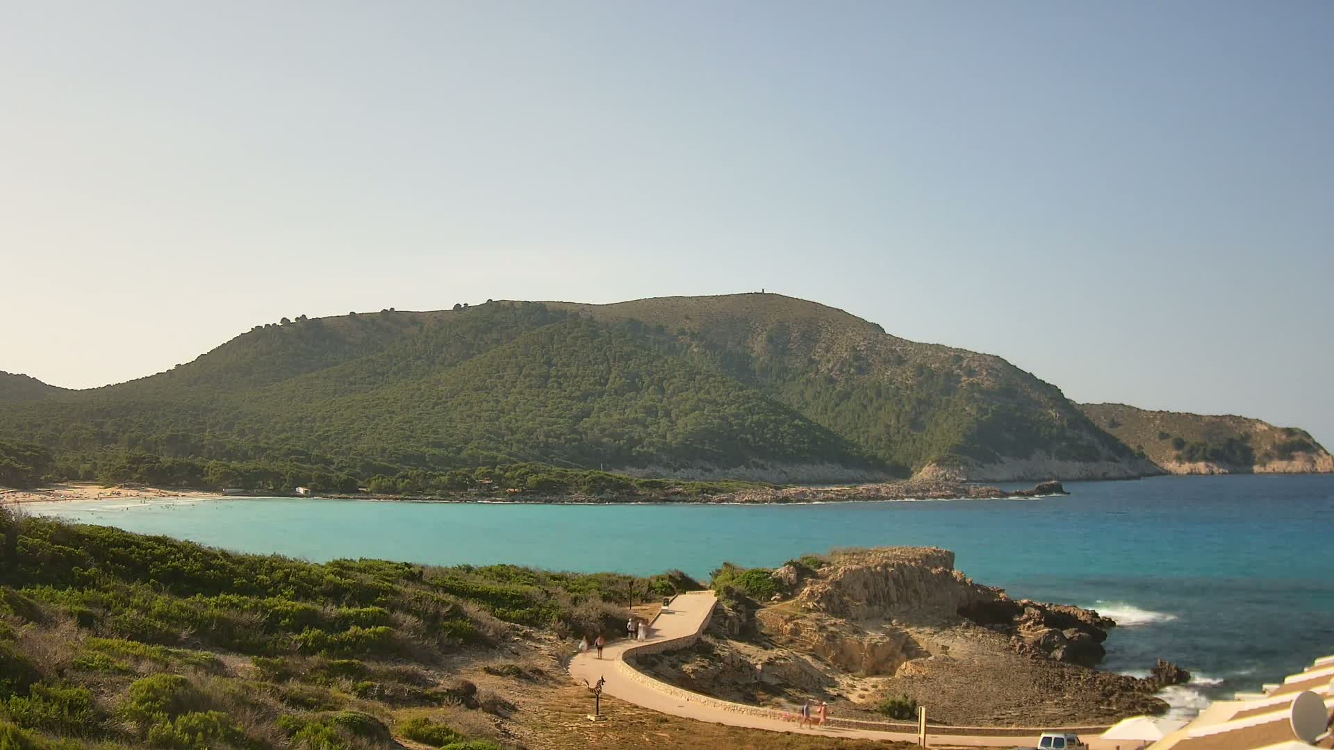 A curving path leads to a secluded turquoise cove nestled beneath a verdant, tree-covered hill on a sunny day.