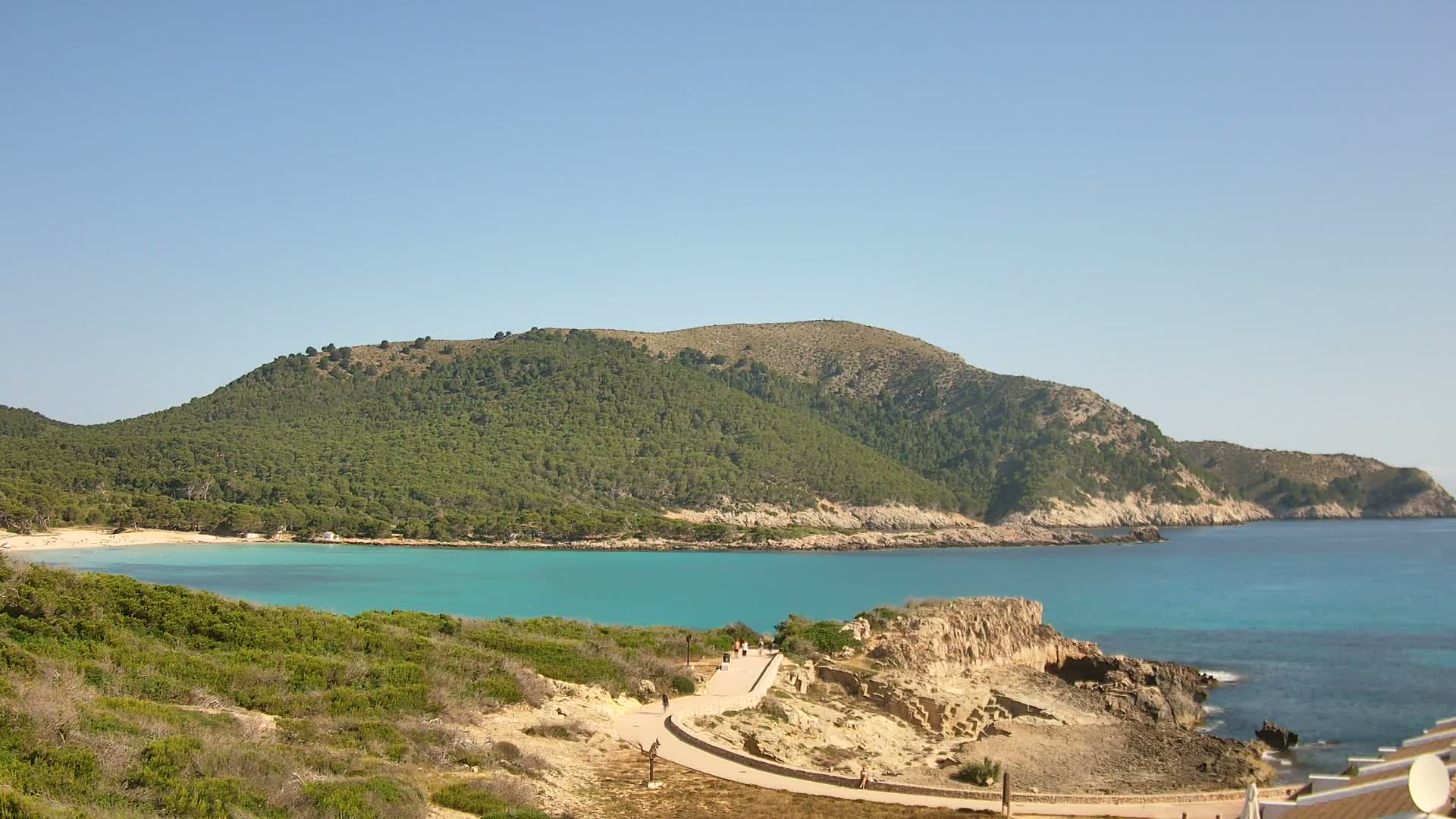 A tranquil cove with turquoise water, a sandy beach, and a verdant, hilly landscape under a clear blue sky.