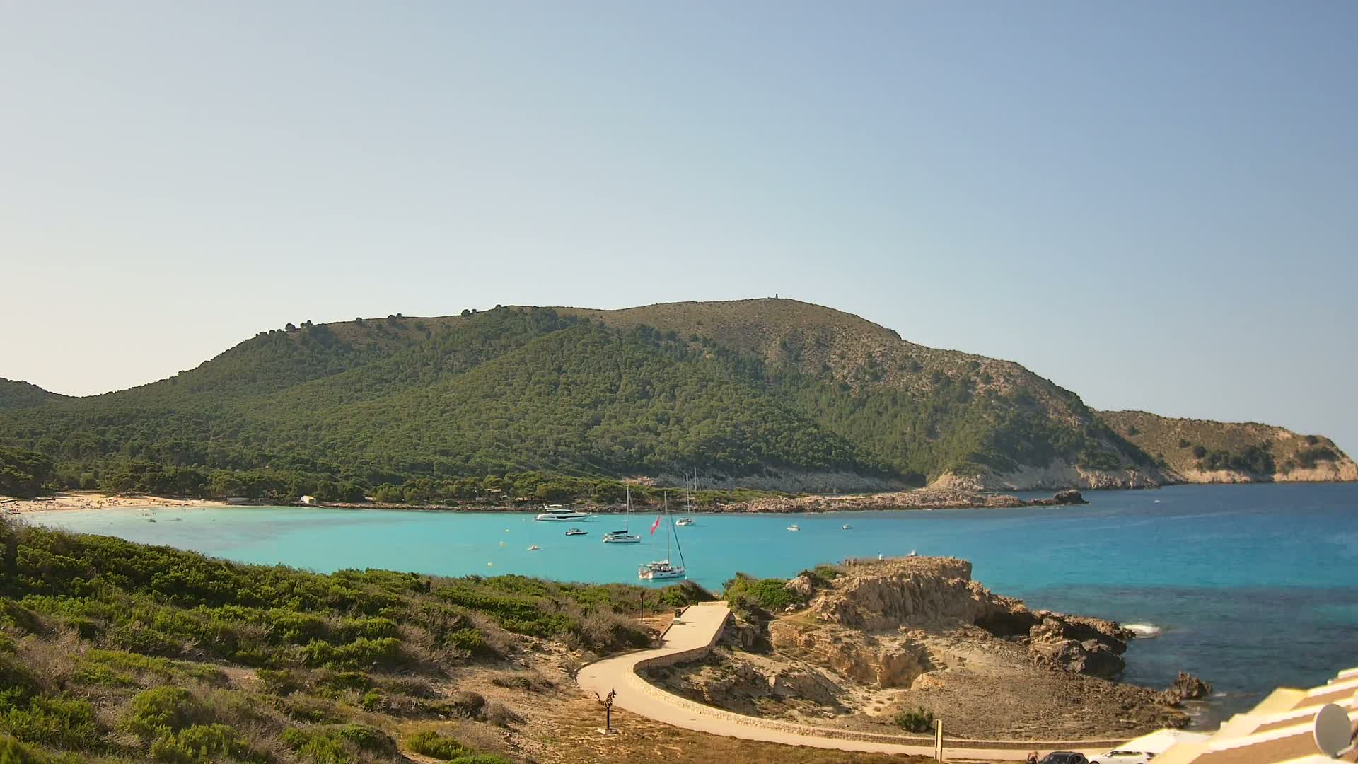 A winding path leads to a turquoise cove with several boats anchored, nestled between a green hill and a rocky shore under a clear sky.