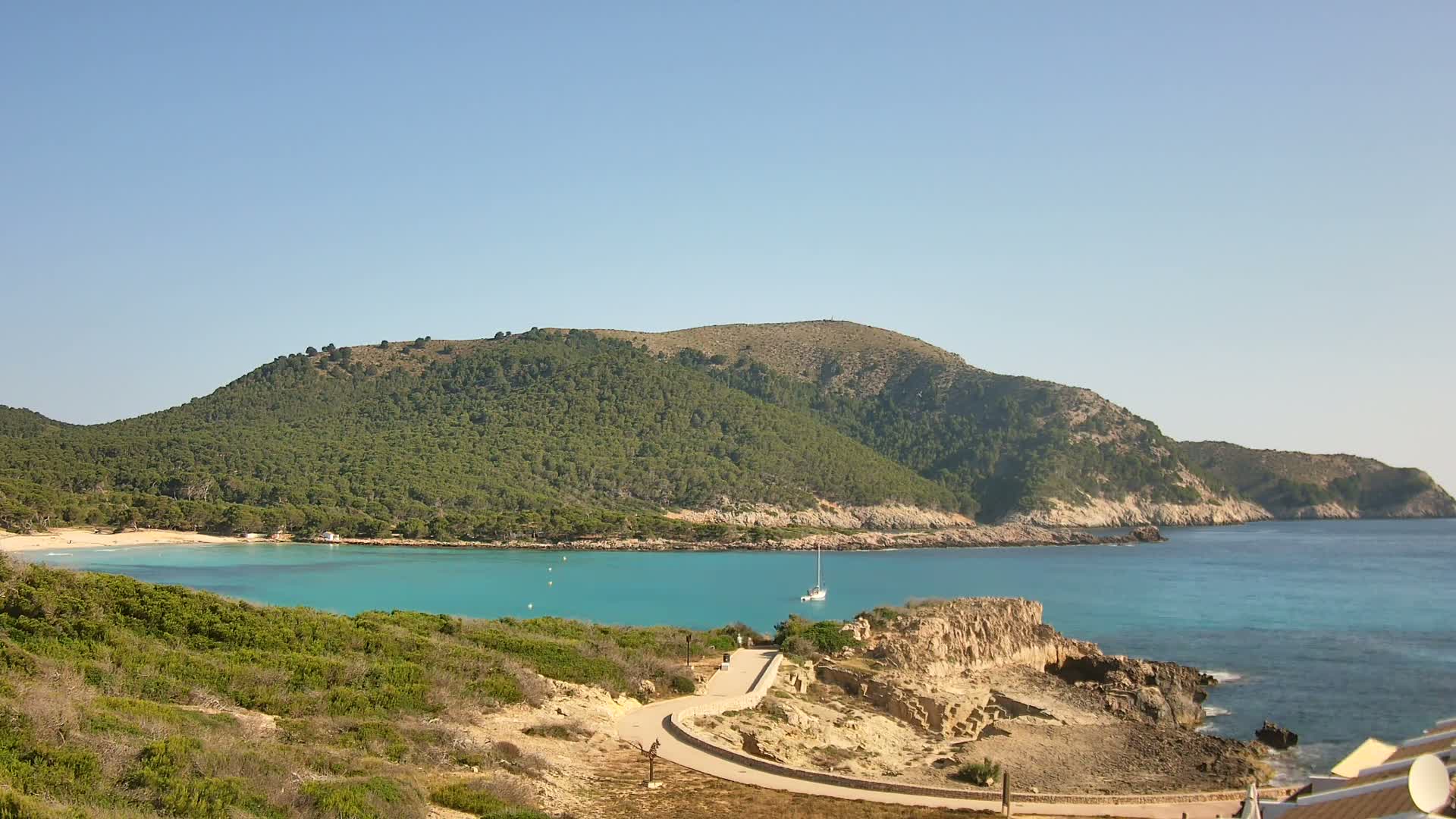 A secluded cove with turquoise water is nestled between a green, mountainous coastline under a clear blue sky.