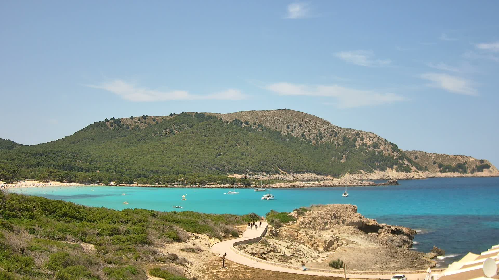 A paved path winds along a rocky coastline overlooking a turquoise bay with a sandy beach and several boats, under a mostly clear blue sky.