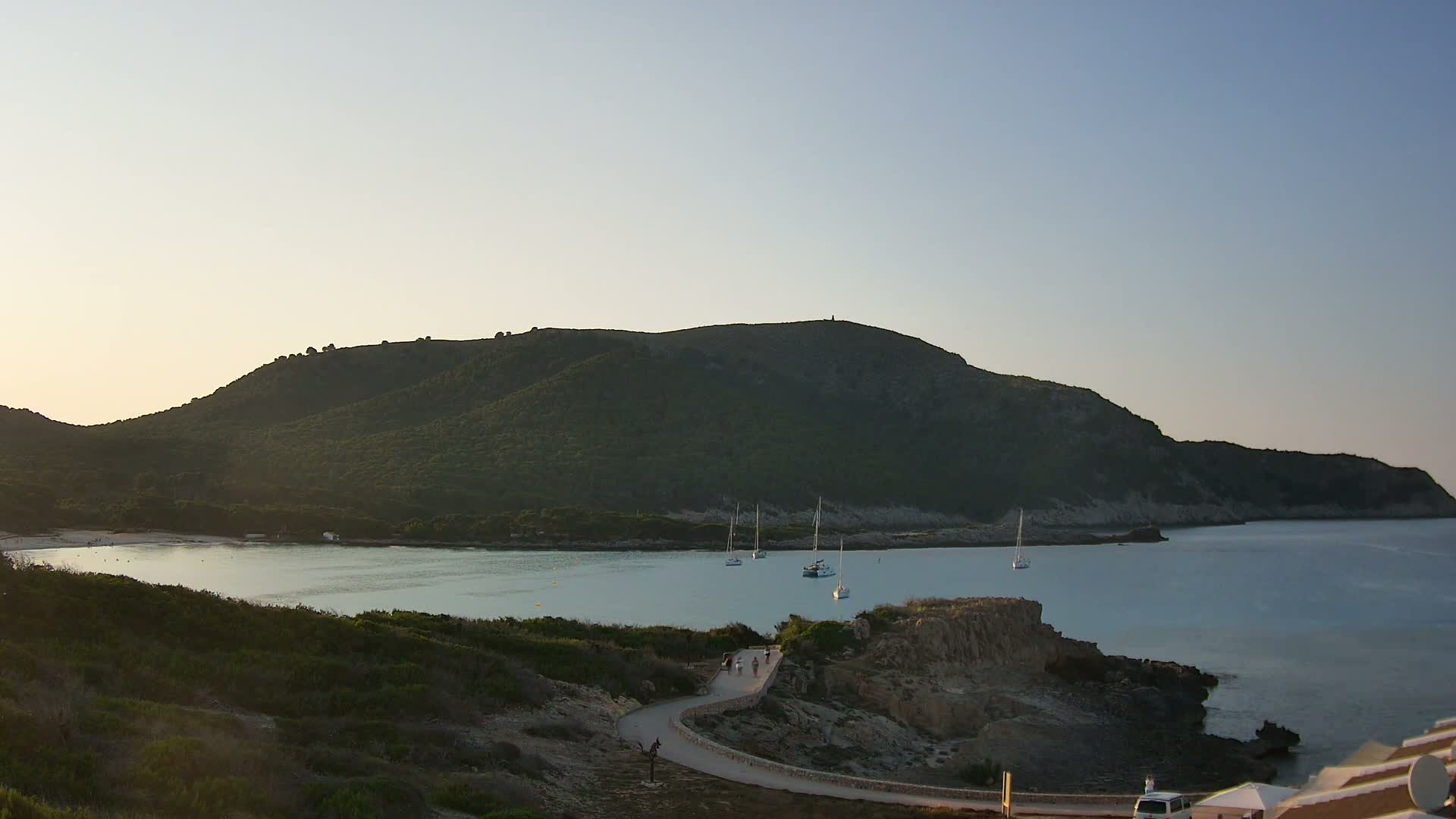 A tranquil bay nestled beneath a verdant hill, with several sailboats anchored in calm, clear water under a clear, light-blue sky.