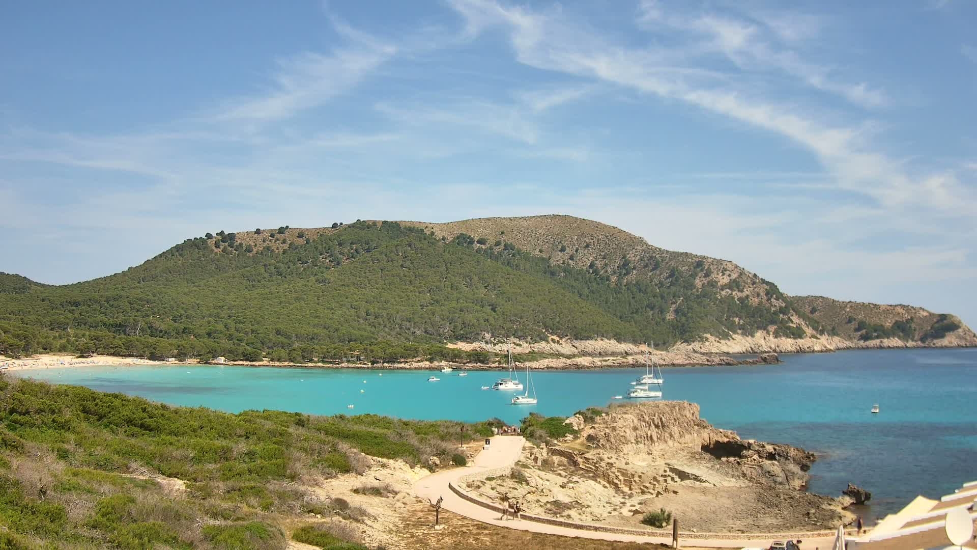 A sunny day reveals a secluded cove with turquoise water, several boats anchored in the bay, and a green, hilly landscape under a mostly clear sky.