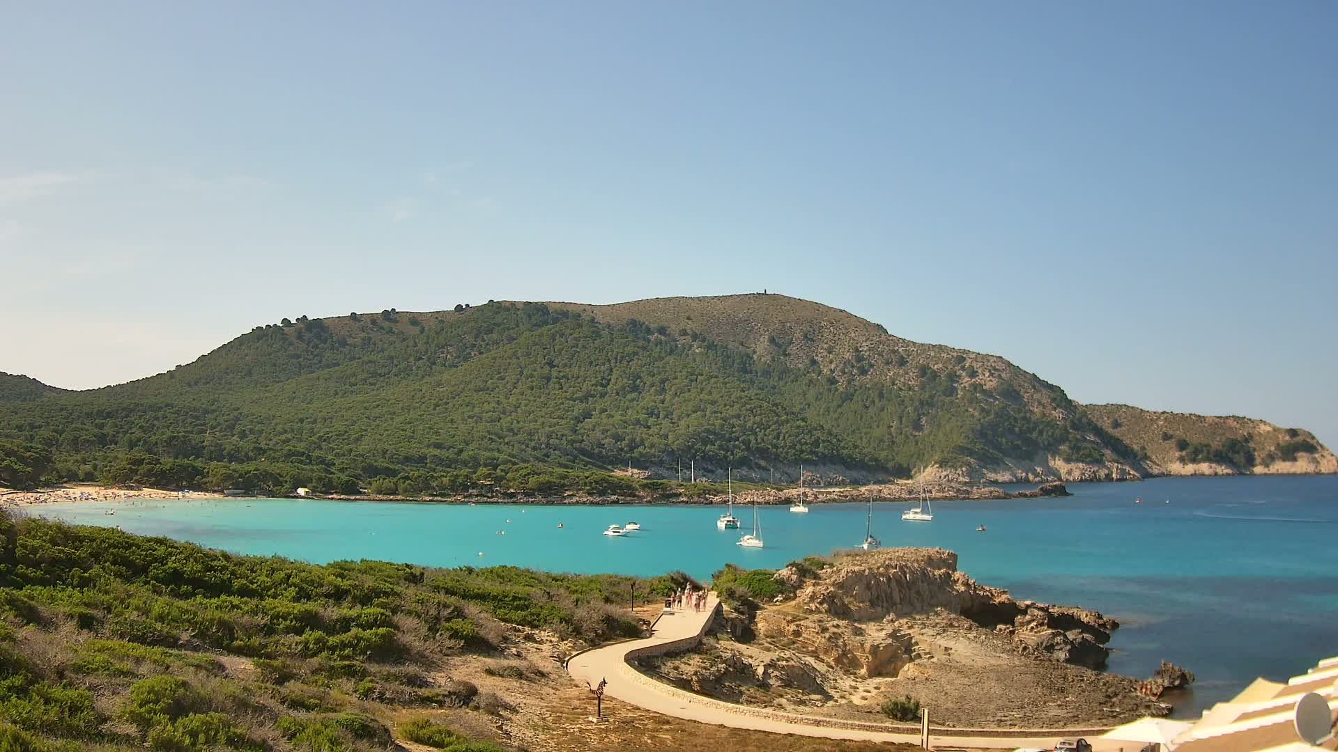 A paved path winds down to a turquoise cove, where several sailboats are anchored near a sandy beach backed by a lush, green hill under a clear, sunny sky.