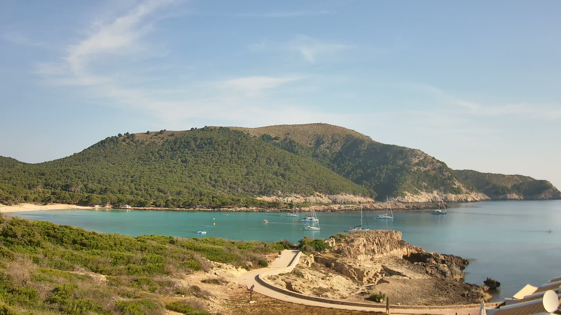A tranquil cove with turquoise water, a sandy beach, and several sailboats anchored near a lush, green, hilly coastline under a mostly sunny sky.