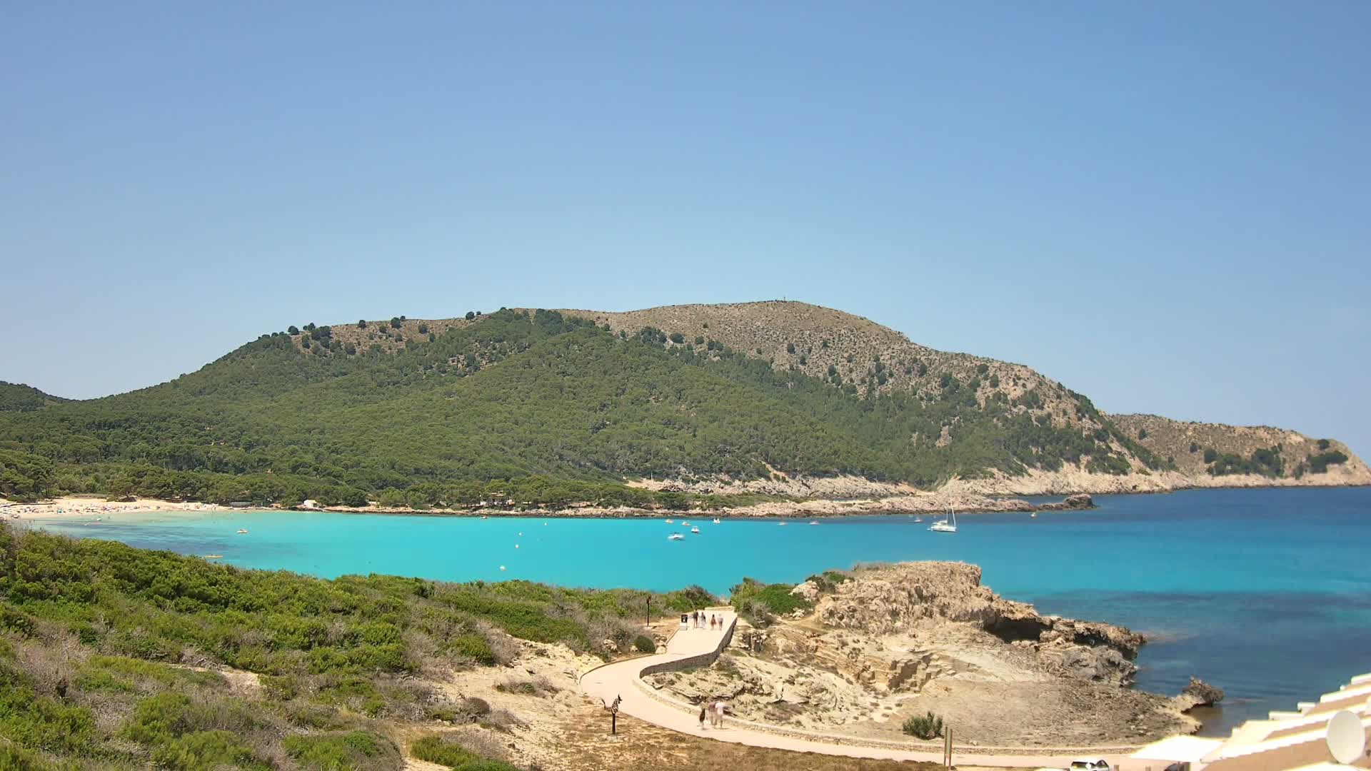 A tranquil turquoise cove nestled between a verdant hillside and a rocky shoreline on a sunny day.