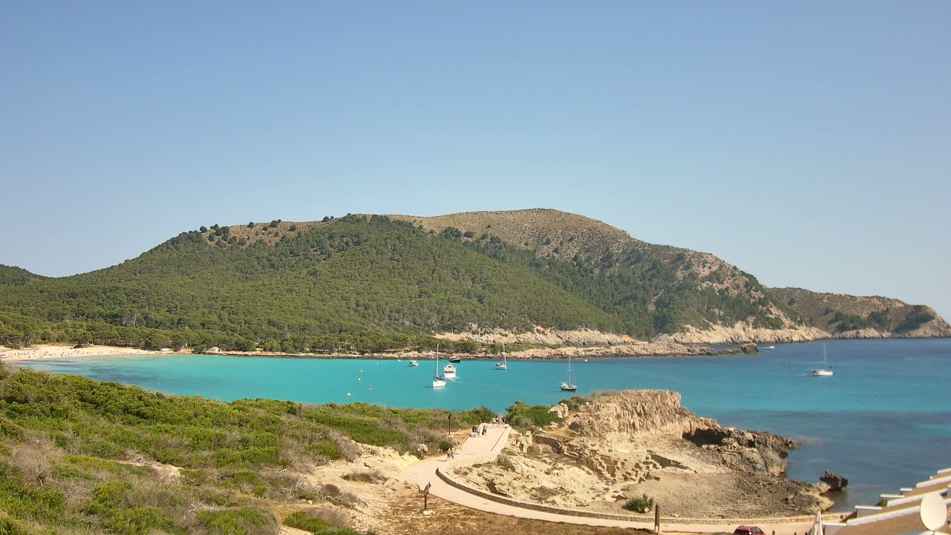 A picturesque cove with turquoise water, a sandy beach, and a verdant hillside under a clear blue sky.