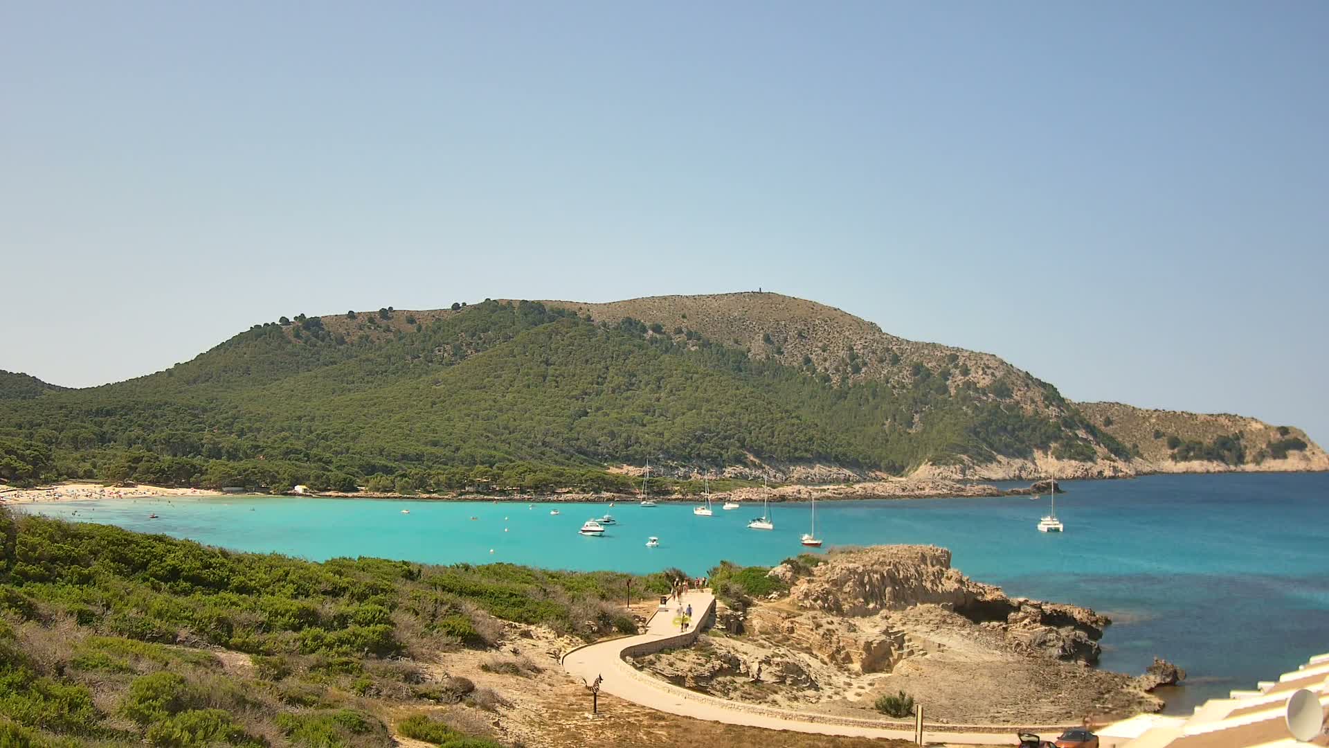 A curving paved path leads to a tranquil turquoise bay with several sailboats anchored, a sandy beach dotted with people, and a verdant, hilly coastline under a clear sky.