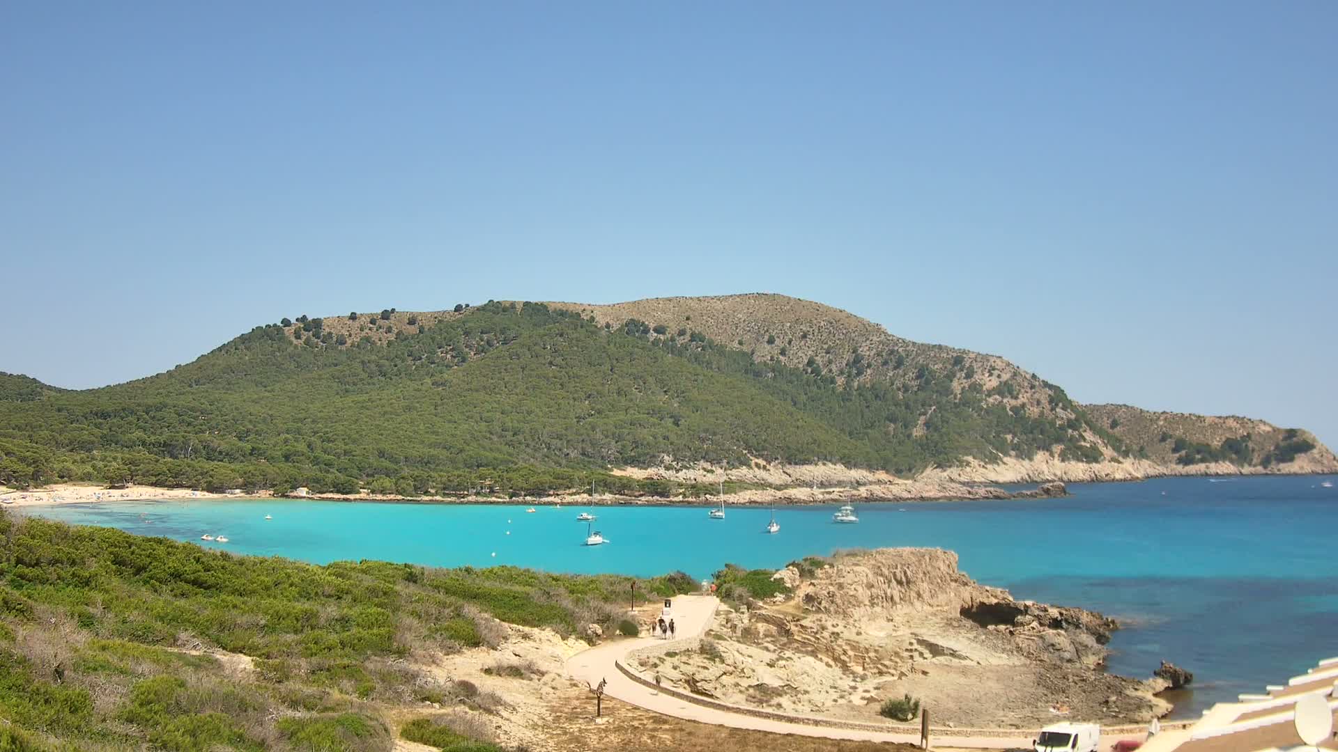 A tranquil cove with turquoise water, a sandy beach, and several boats is nestled against a lush, green hill under a clear blue sky.