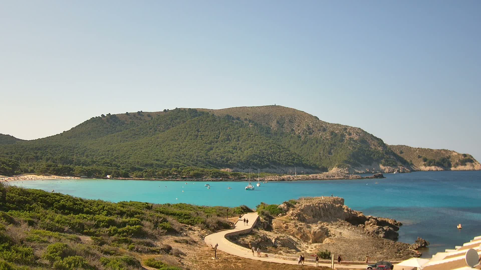 A curving path leads down to a turquoise cove nestled between a verdant hillside and a clear blue sea on a sunny day.