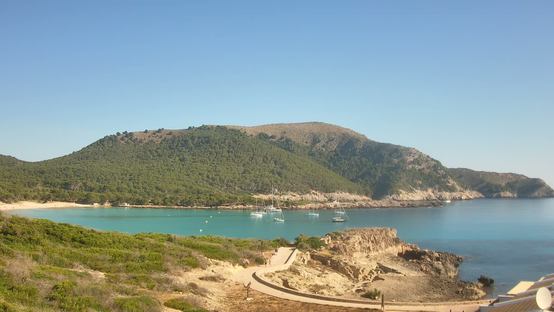 A tranquil turquoise bay is nestled within a verdant, hilly coastline under a clear blue sky.