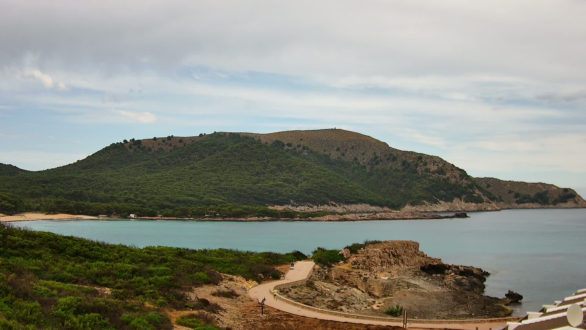 A winding road leads to a tranquil turquoise bay nestled between a lush, green hill and a rocky coastline under a mostly cloudy sky.
