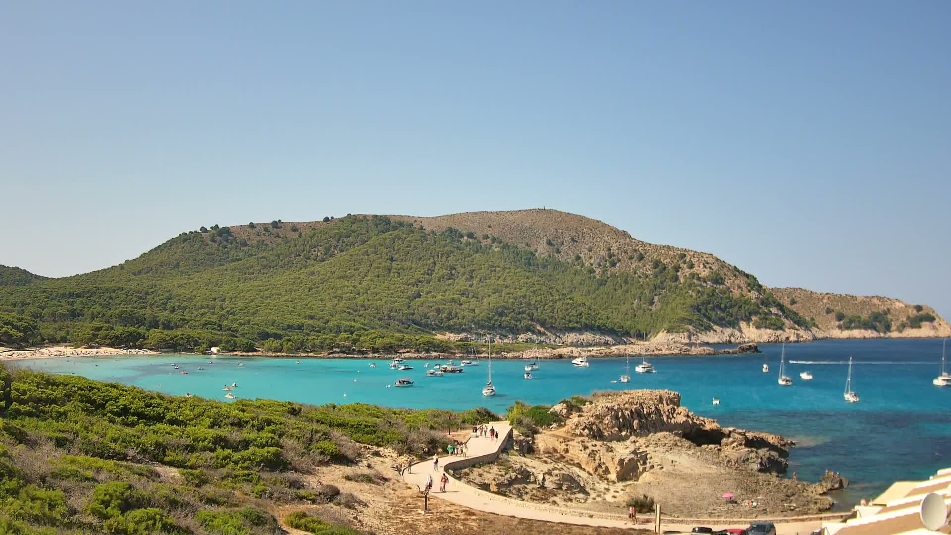 A tranquil cove with turquoise water is nestled beneath a verdant hill on a clear, sunny day, and two sailboats are anchored in the bay.