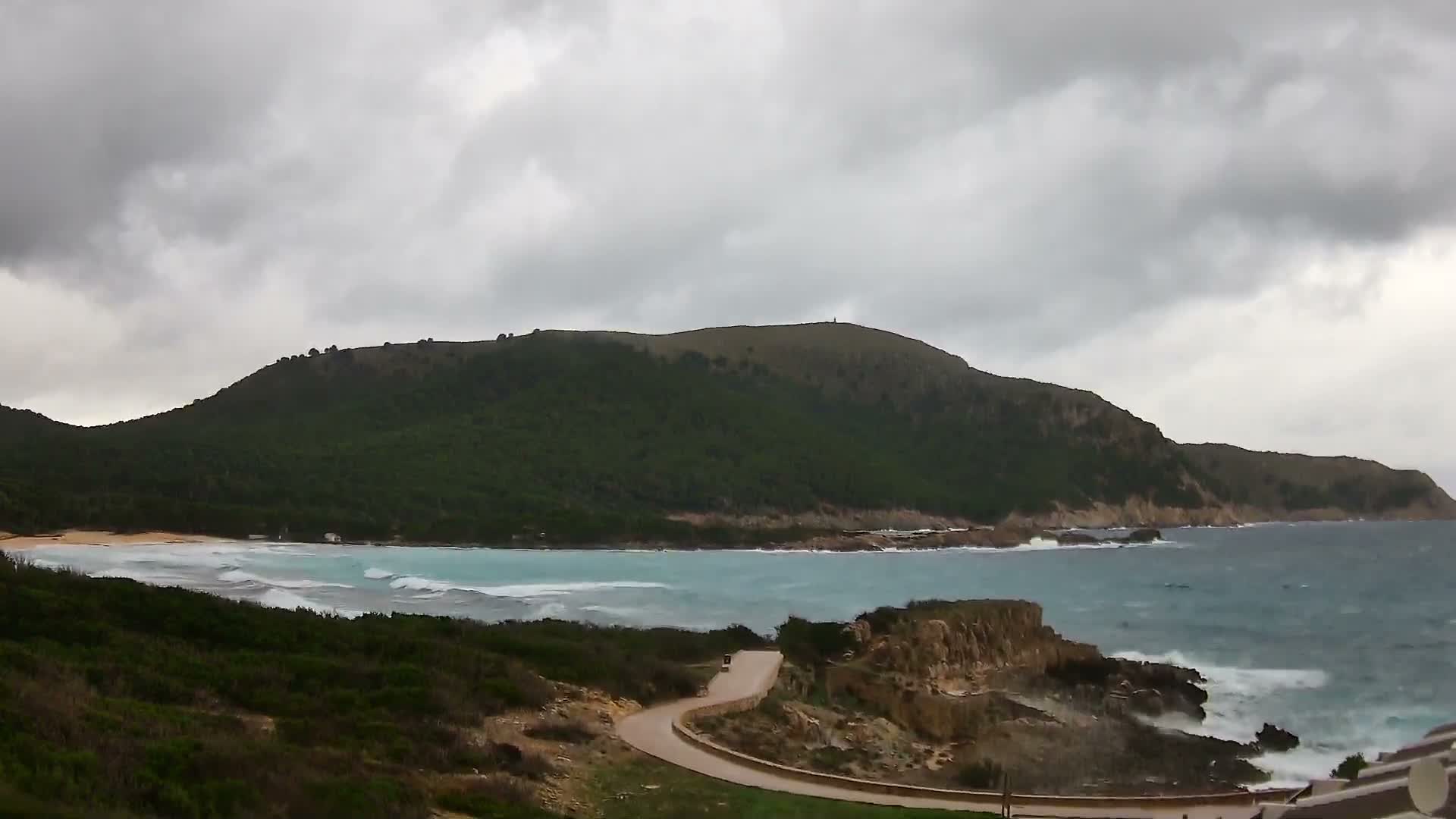 Under a heavily overcast sky, a dramatic coastal landscape features a sweeping green hillside, a sandy bay with powerful turquoise waves, and a winding road along rocky cliffs meeting the rough sea.