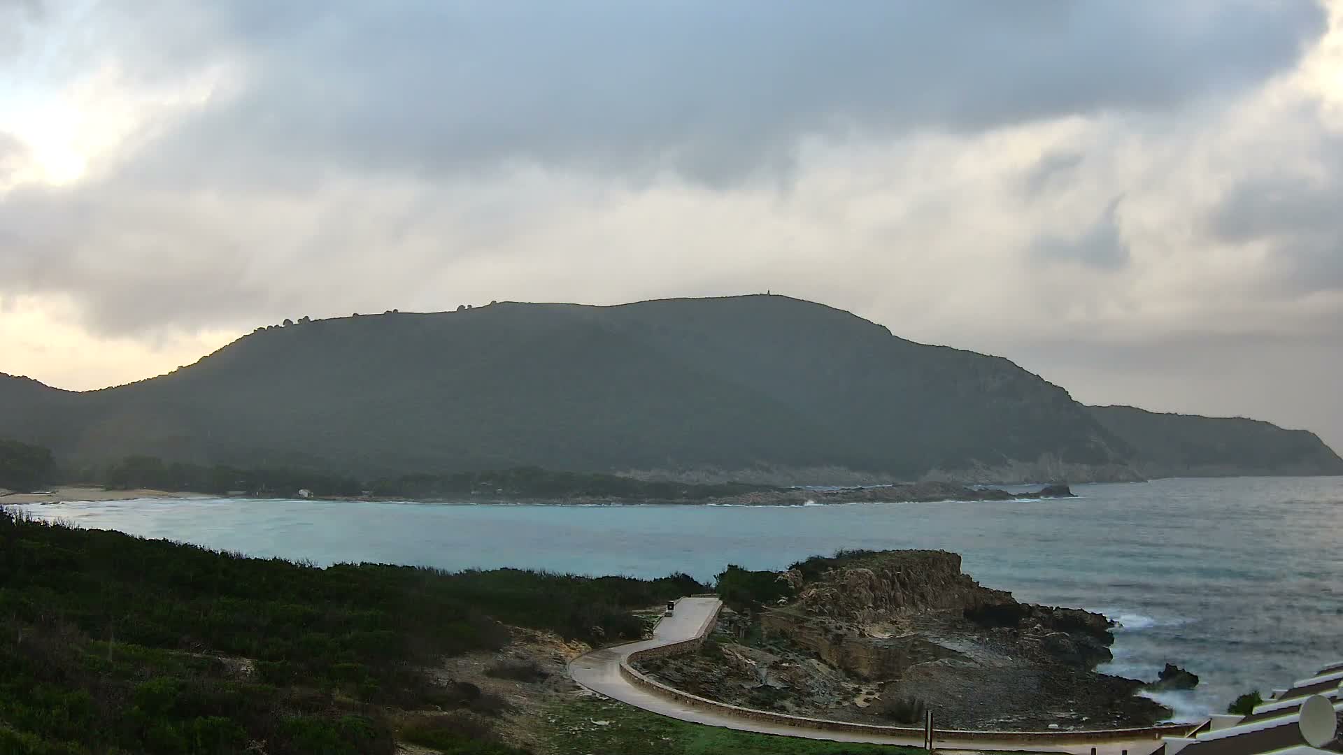 On an overcast day, a winding coastal path overlooks rocky shores with breaking waves, a sandy cove, and verdant hills in the distance.