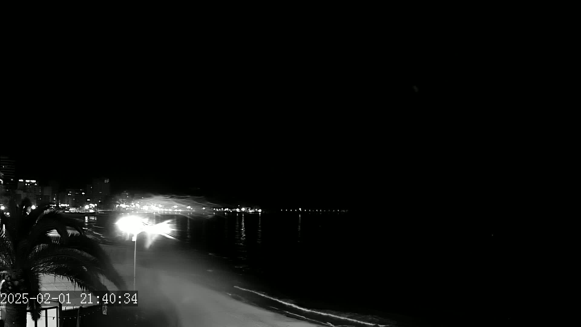 A nighttime, black and white view of a beach with city lights across the water, showing a windy or stormy condition.