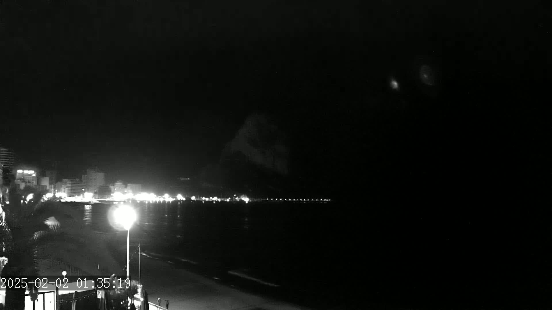 A nighttime, black and white view of a dark ocean with a brightly lit city skyline in the distance.