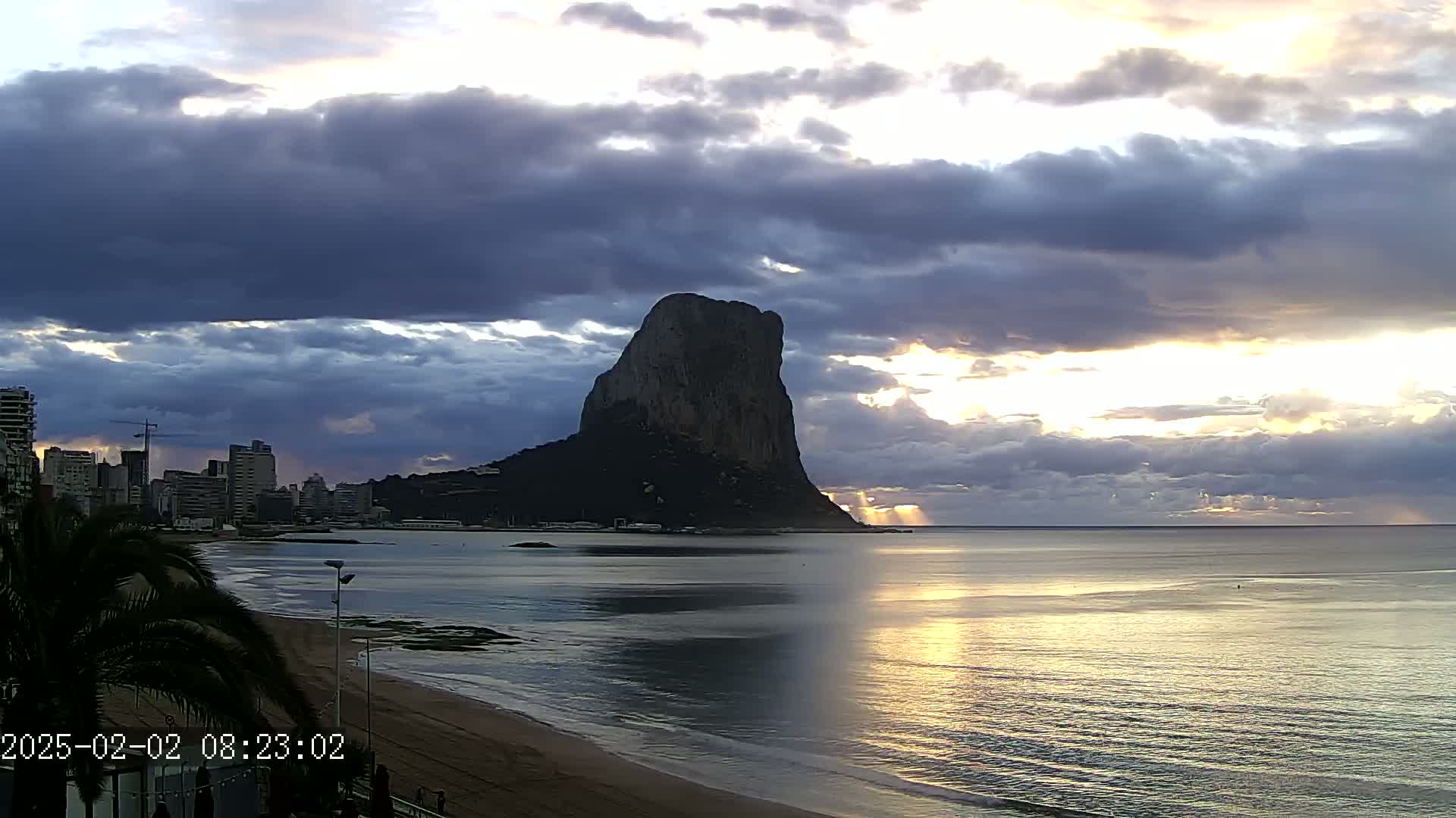 A large, dark rock formation rises from the sea under a partly cloudy sunrise sky, with a city skyline visible to the left and a sandy beach in the foreground.