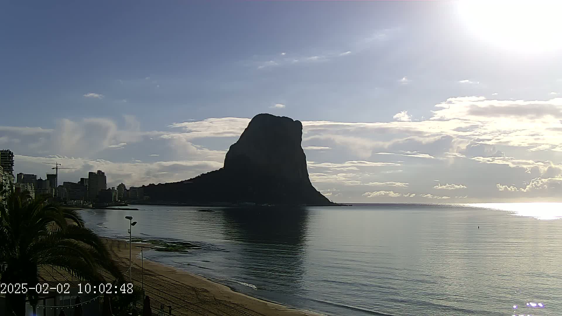 A large, dark rock formation rises from the calm, sunlit sea near a sandy beach and a city skyline under a partly cloudy sky.