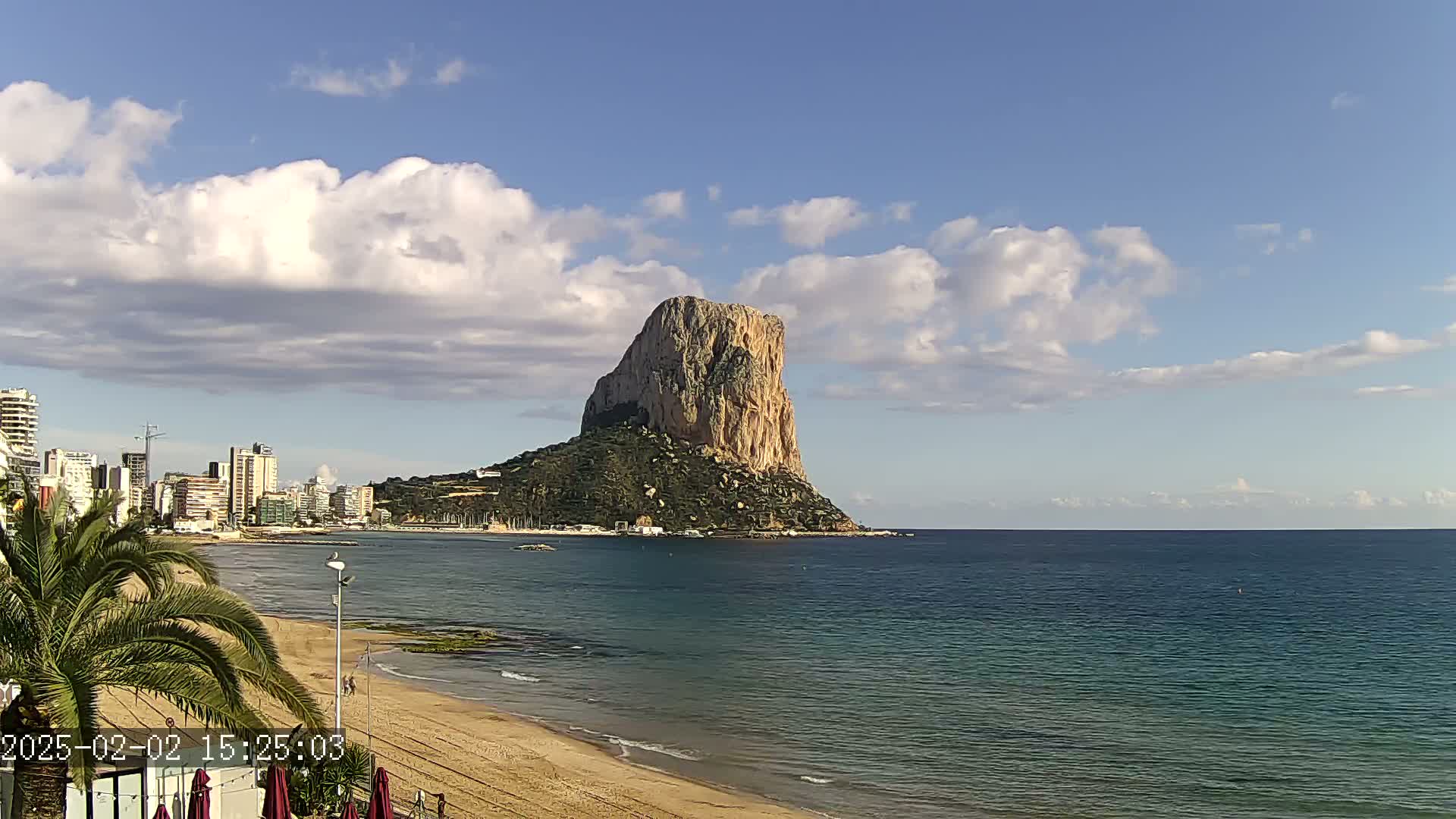 A large, rocky island rises from the sea under a partly cloudy sky, with a sandy beach and a city skyline visible in the background.