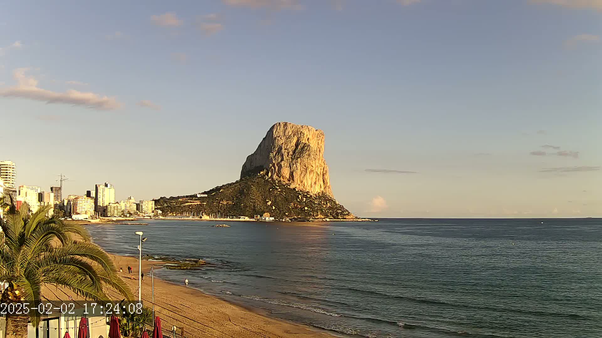 A large, tan rock formation rises from the sea near a sandy beach and a city skyline under a mostly clear, light-blue sky.