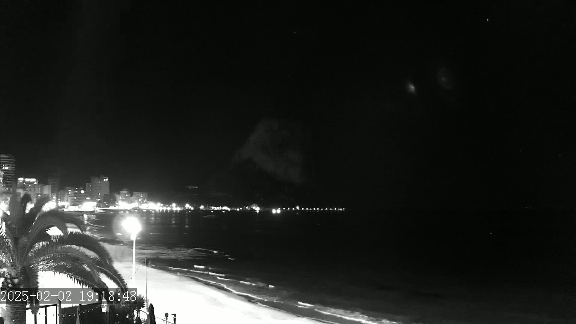 A nighttime, black and white view of a beach with a city skyline in the distance, under a clear sky with some faint, bright objects visible.
