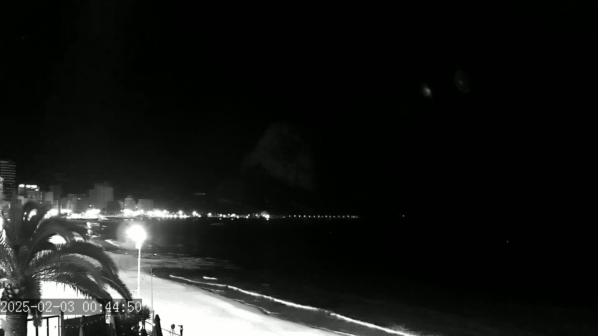 A nighttime view of a beach with a city skyline and a dark, mountainous silhouette in the background under a clear sky.