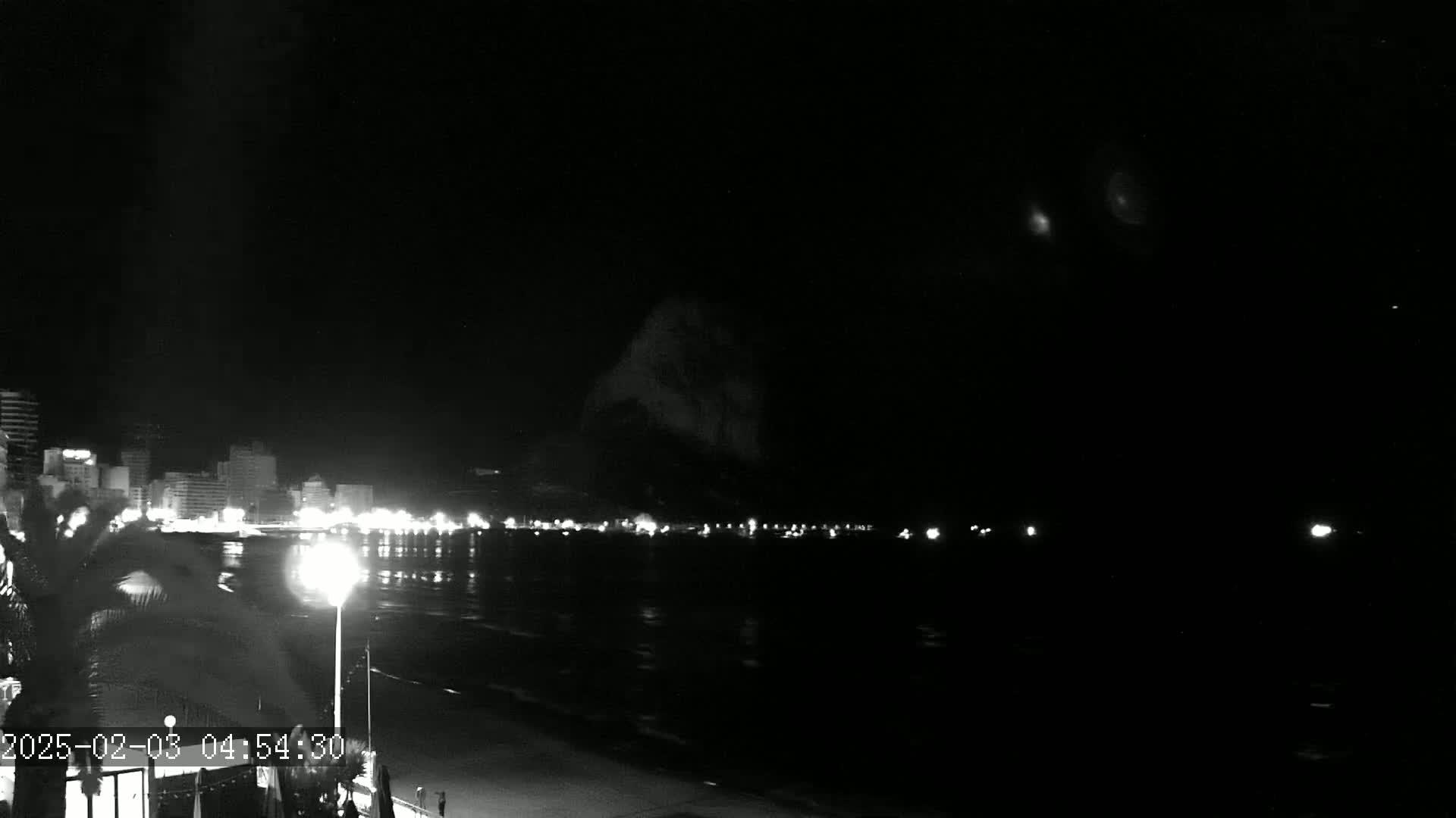 A nighttime, seaside view shows a dark, calm body of water reflecting the lights of a distant city skyline under a mostly clear sky.