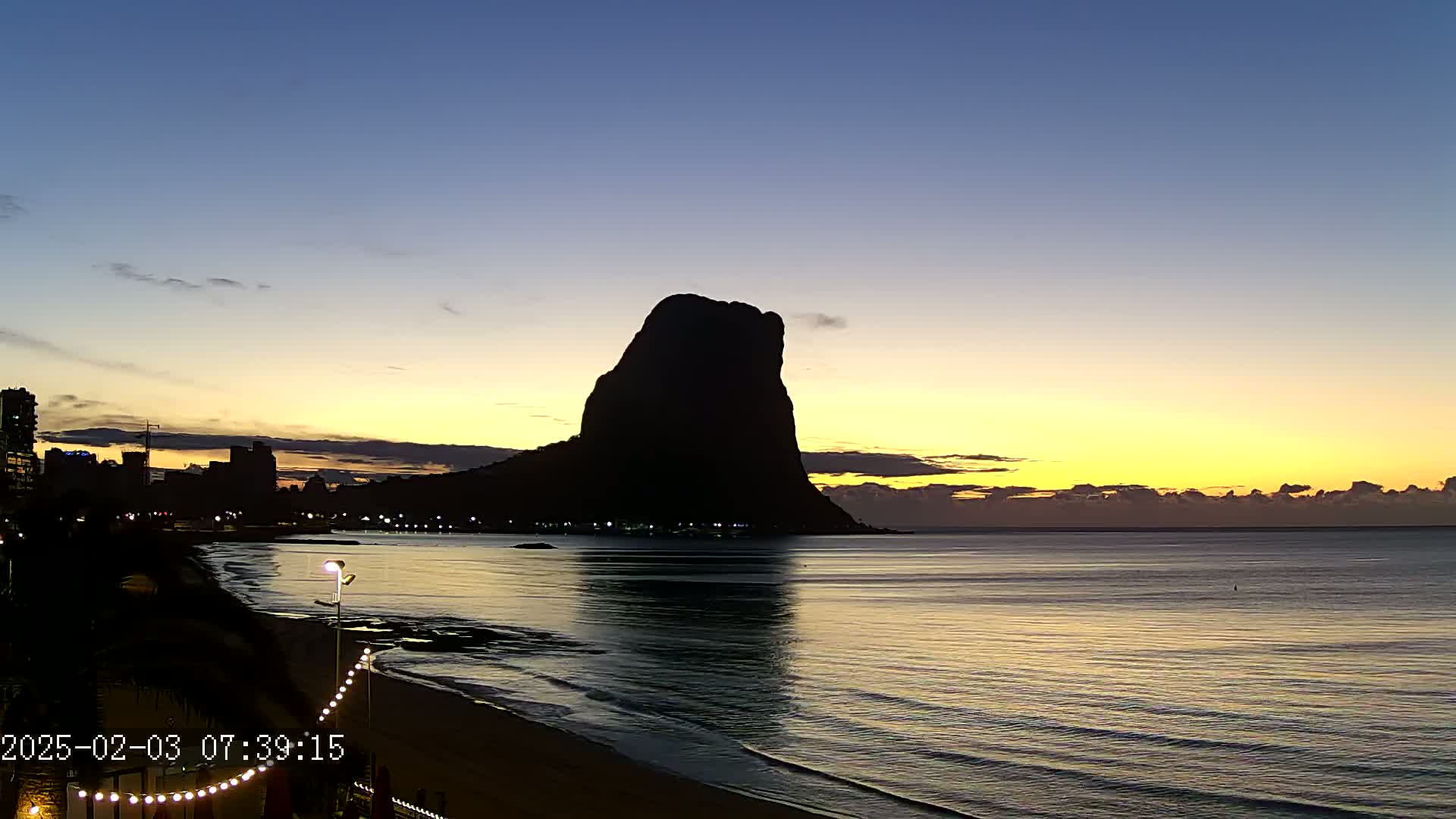 A large, dark rock formation silhouetted against a sunrise over a calm ocean, with a city skyline visible in the distance.