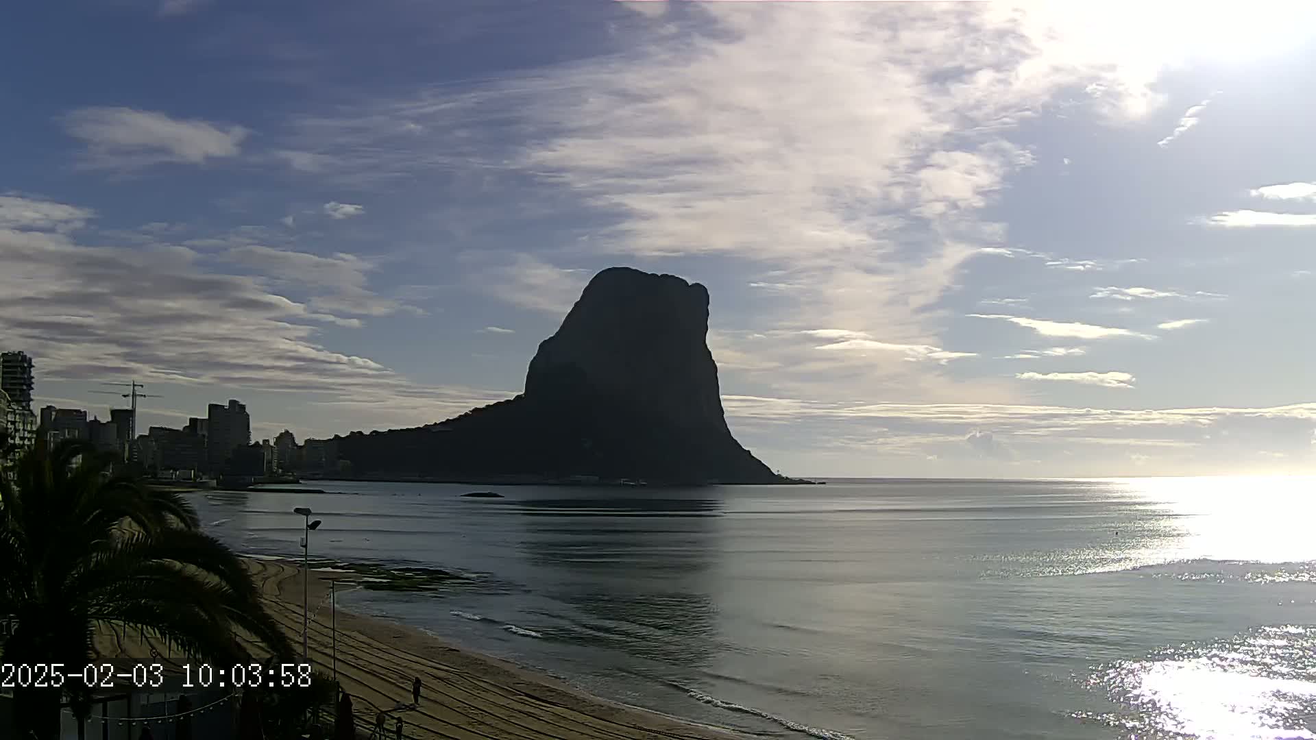 A large, dark, solitary mountain rises from the ocean under a partly cloudy sky, with a sandy beach and a city skyline visible in the foreground.
