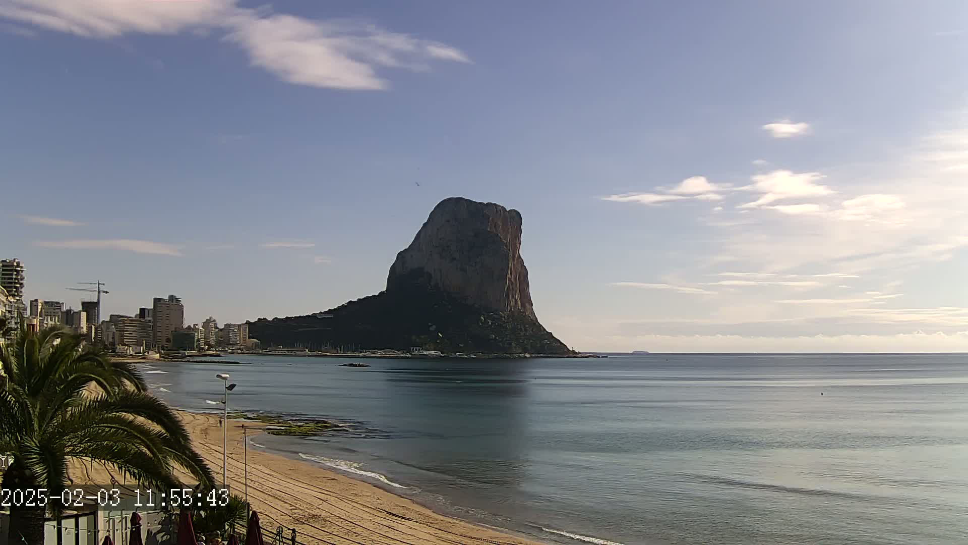 A large, rocky island rises from the calm sea, a sandy beach and a city skyline visible in the background under a partly cloudy sky.