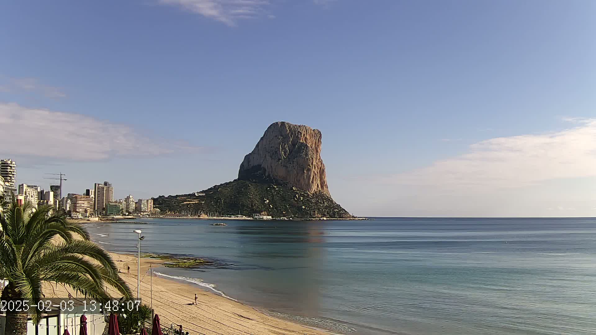 A large, rocky island rises from the calm, blue sea, with a sandy beach and cityscape visible in the foreground under a mostly clear sky.