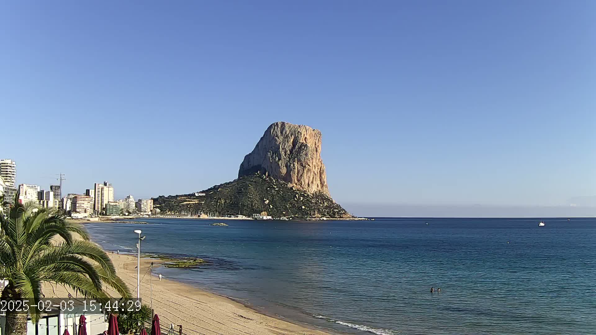A large, rocky island rises from a calm, blue sea next to a sandy beach and a cityscape under a clear blue sky.