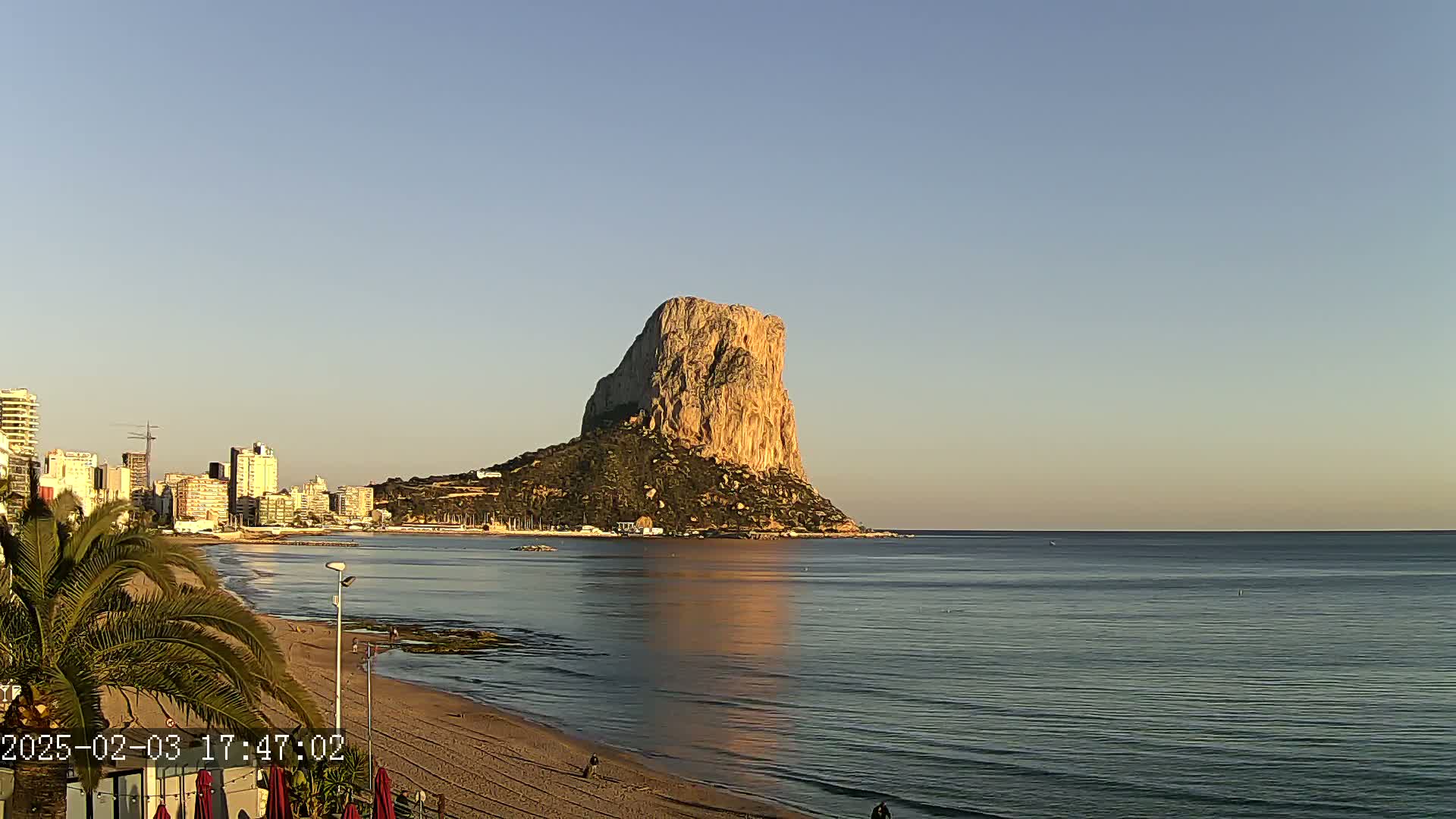 A large, tan rock formation rises from the sea near a sandy beach and a city skyline under a clear, light-blue sky.
