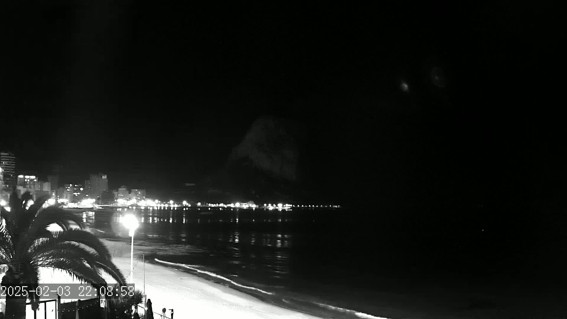 A nighttime, black-and-white view of a beach at night, showing a calm sea reflecting the lights of buildings on the shore and a dark mountain in the background.