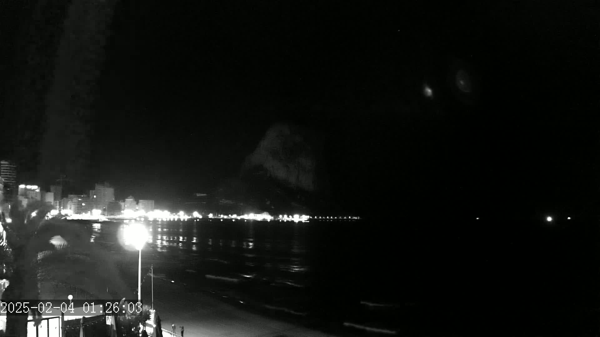 A nighttime, seaside view shows a dark beach and ocean under a clear sky, with a line of illuminated buildings along the shore.