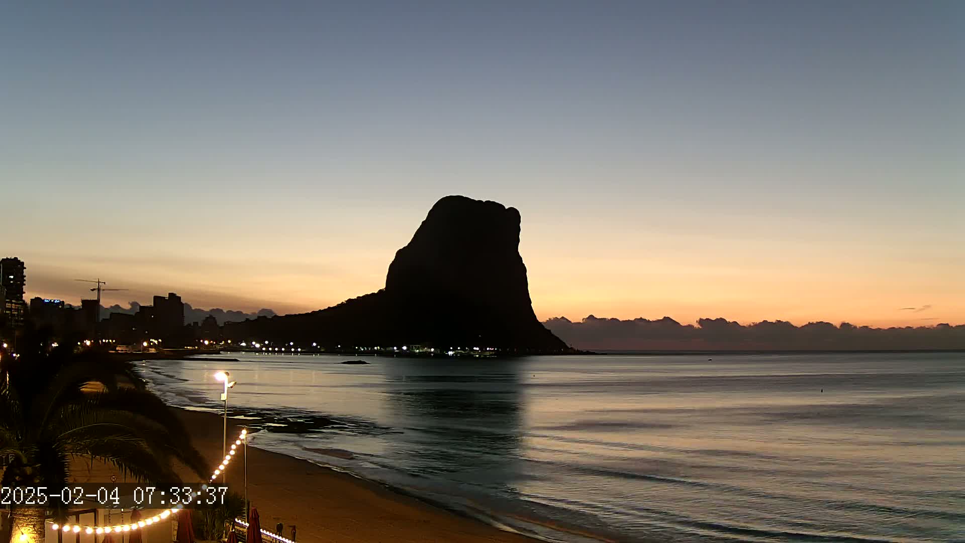 A large, dark silhouette of a rock formation rises from a calm ocean at sunrise, with a city skyline visible in the distance.