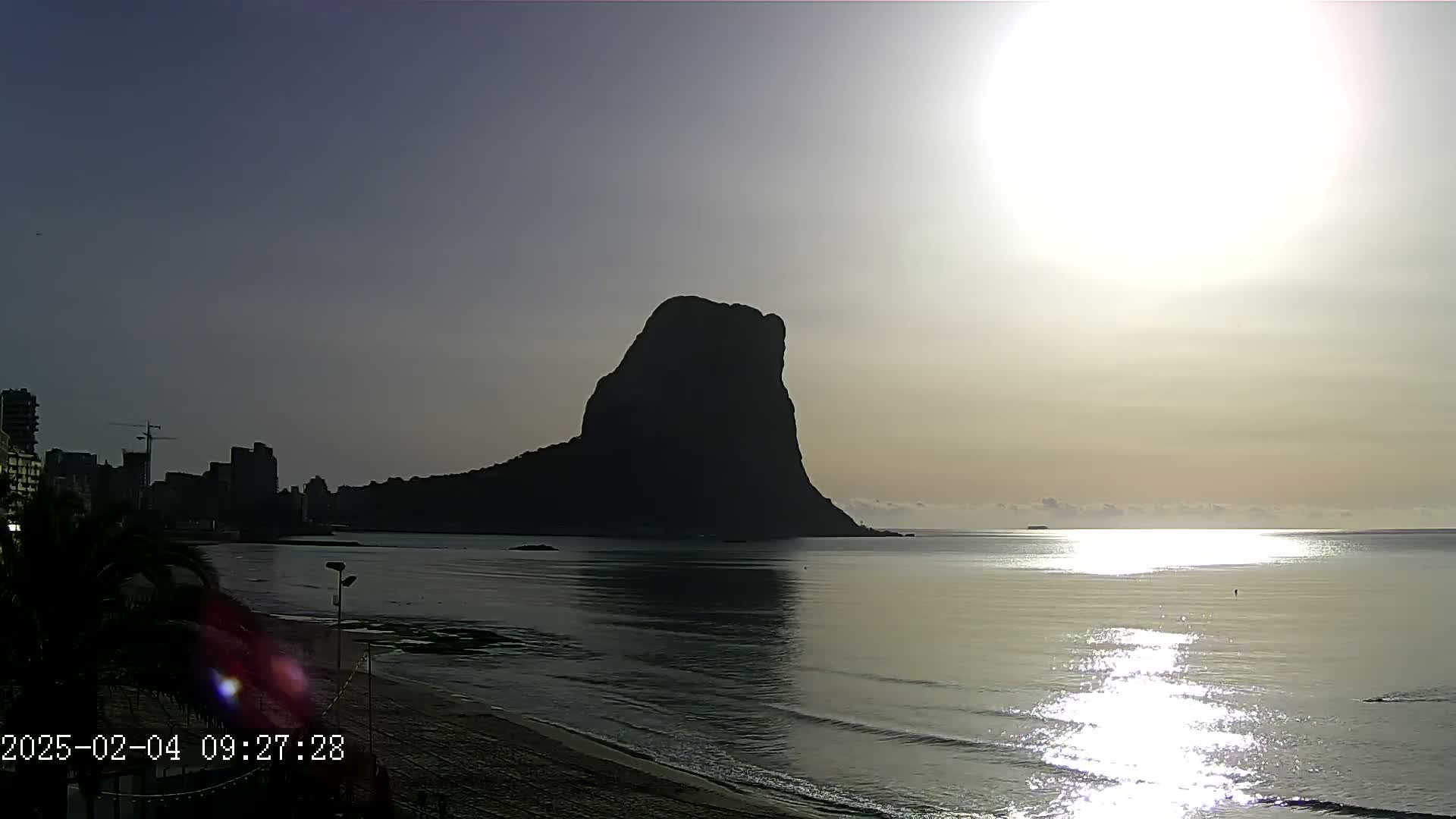 A large, dark, solitary rock formation stands out against a bright, sunrise sky over a calm, reflective sea near a city skyline.