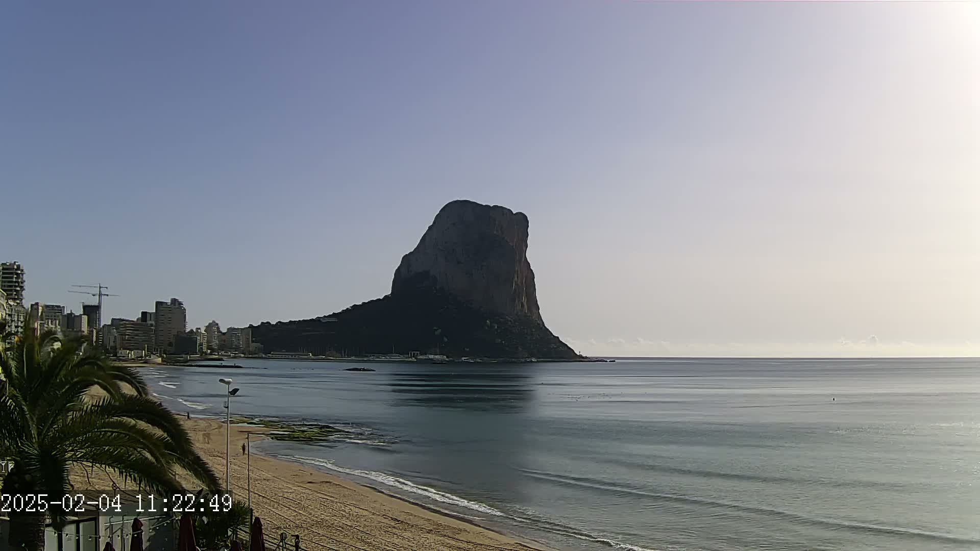 A large, dark rock formation rises from the sea under a clear, light-blue sky, with a sandy beach and a city skyline visible in the foreground.