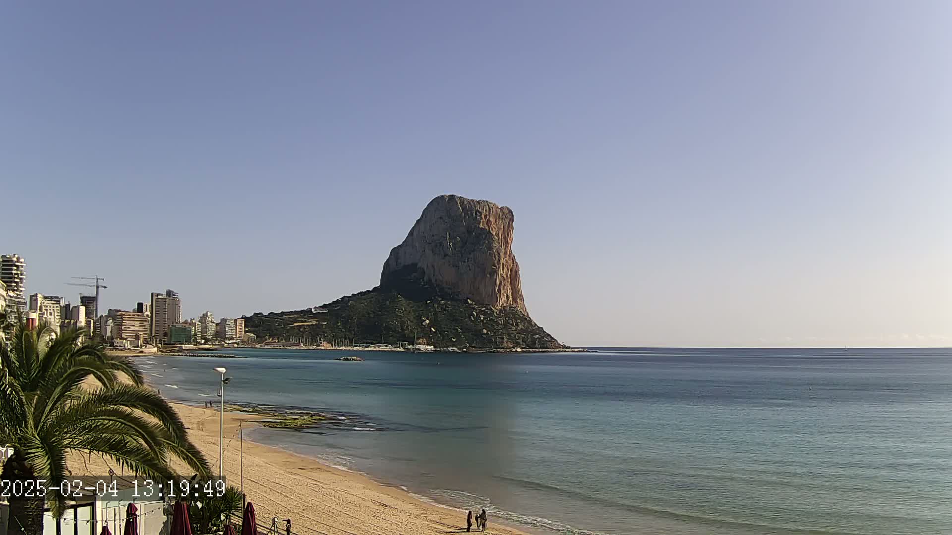 A large, rocky island rises from the calm, blue sea near a sandy beach and a cityscape under a clear, sunny sky.