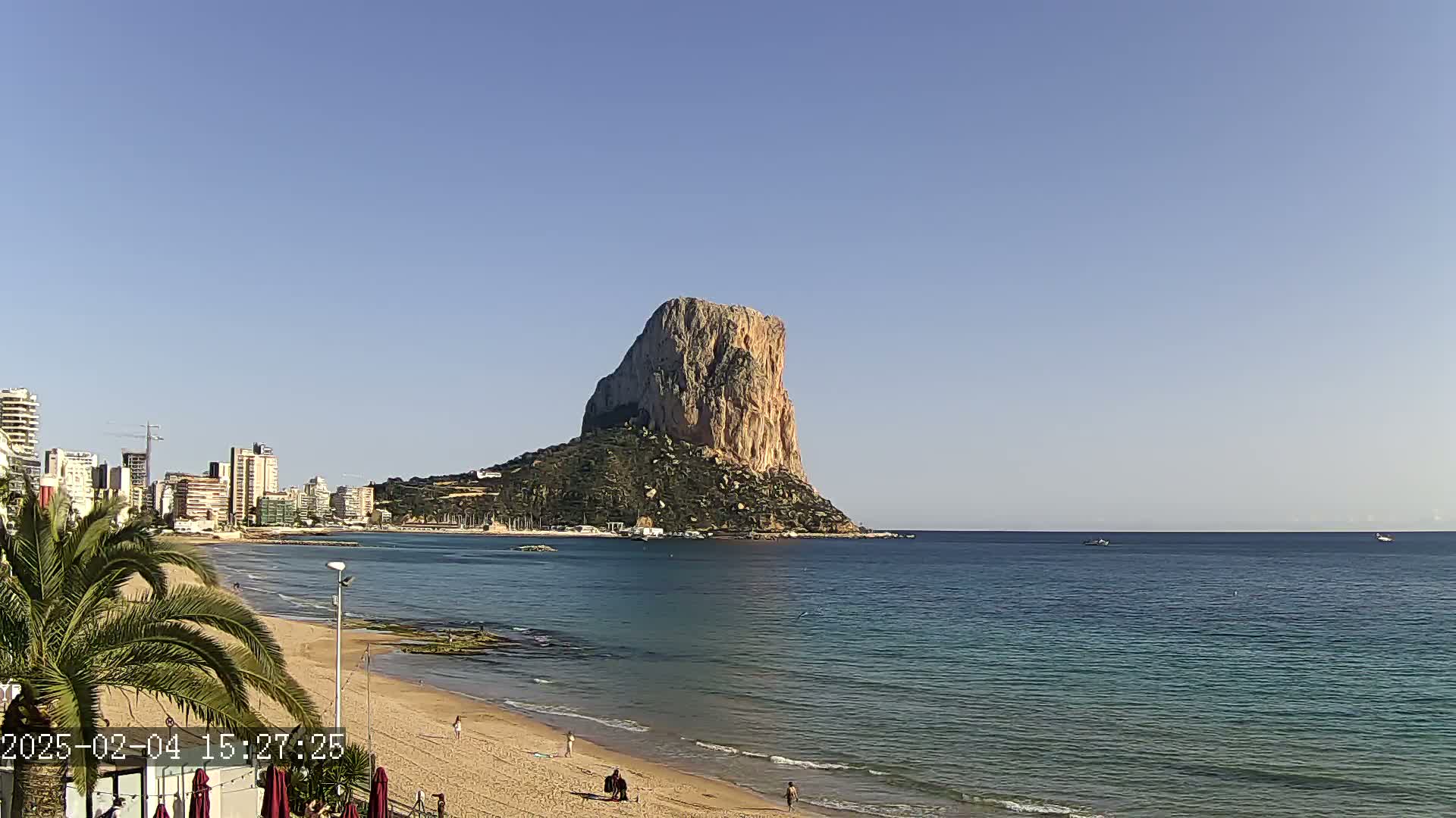 A large, rocky island rises from the calm, teal ocean, with a sandy beach and a city skyline visible in the background under a clear, blue sky.