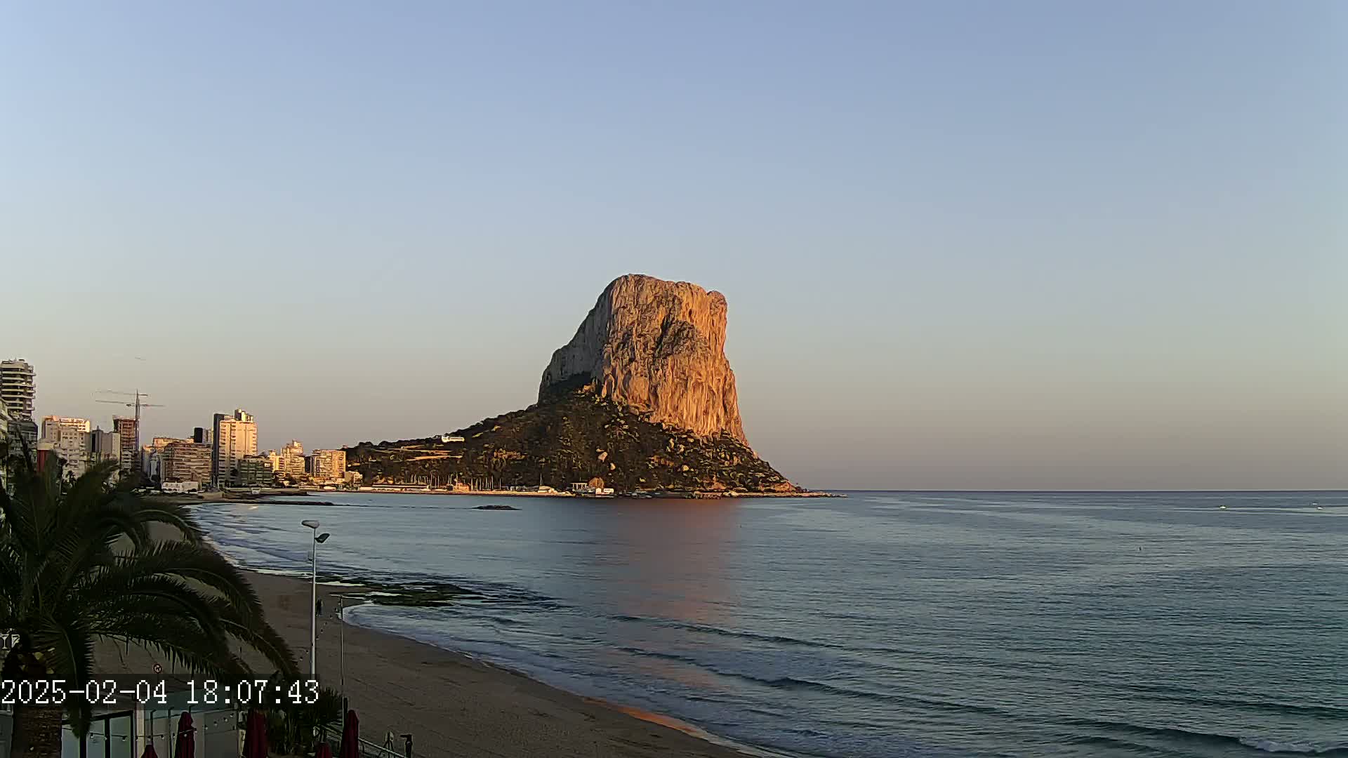 A large, rocky, tan-colored mountain rises from the ocean near a beach and city skyline under a clear, twilight sky.